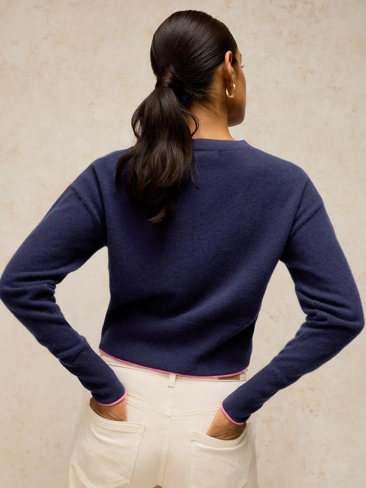 A woman shown from the back wearing a navy blue knit sweater and cream-colored pants, standing against a textured beige wall.