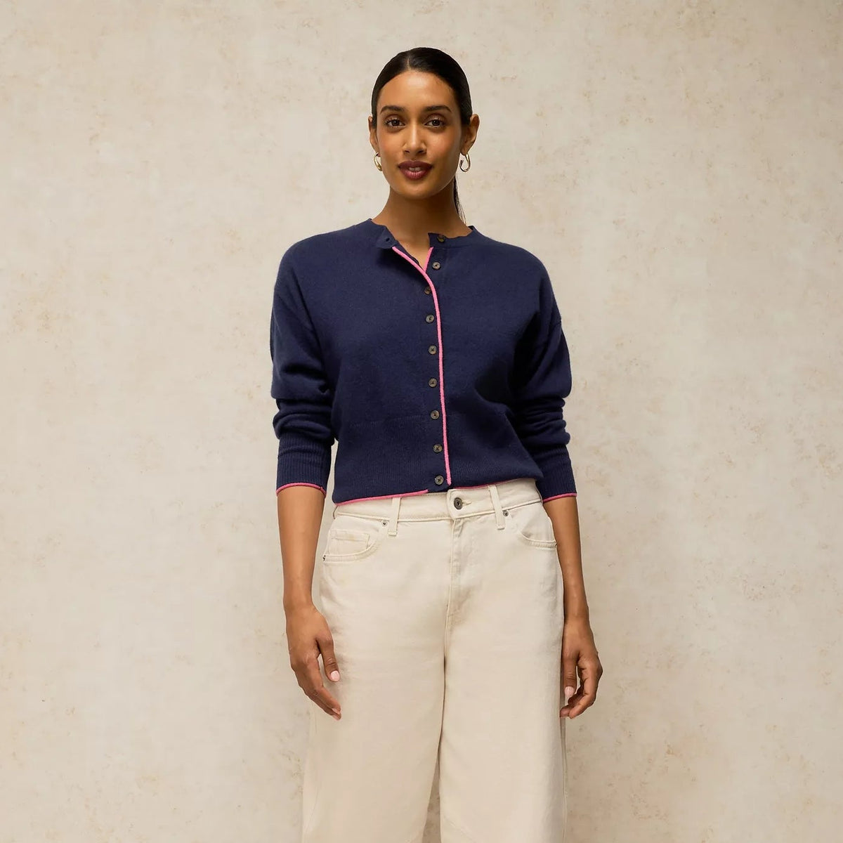 A woman stands against a textured beige studio backdrop wearing a navy cardigan with pink trim over a pink top, cream cropped trousers, and snakeskin flats.