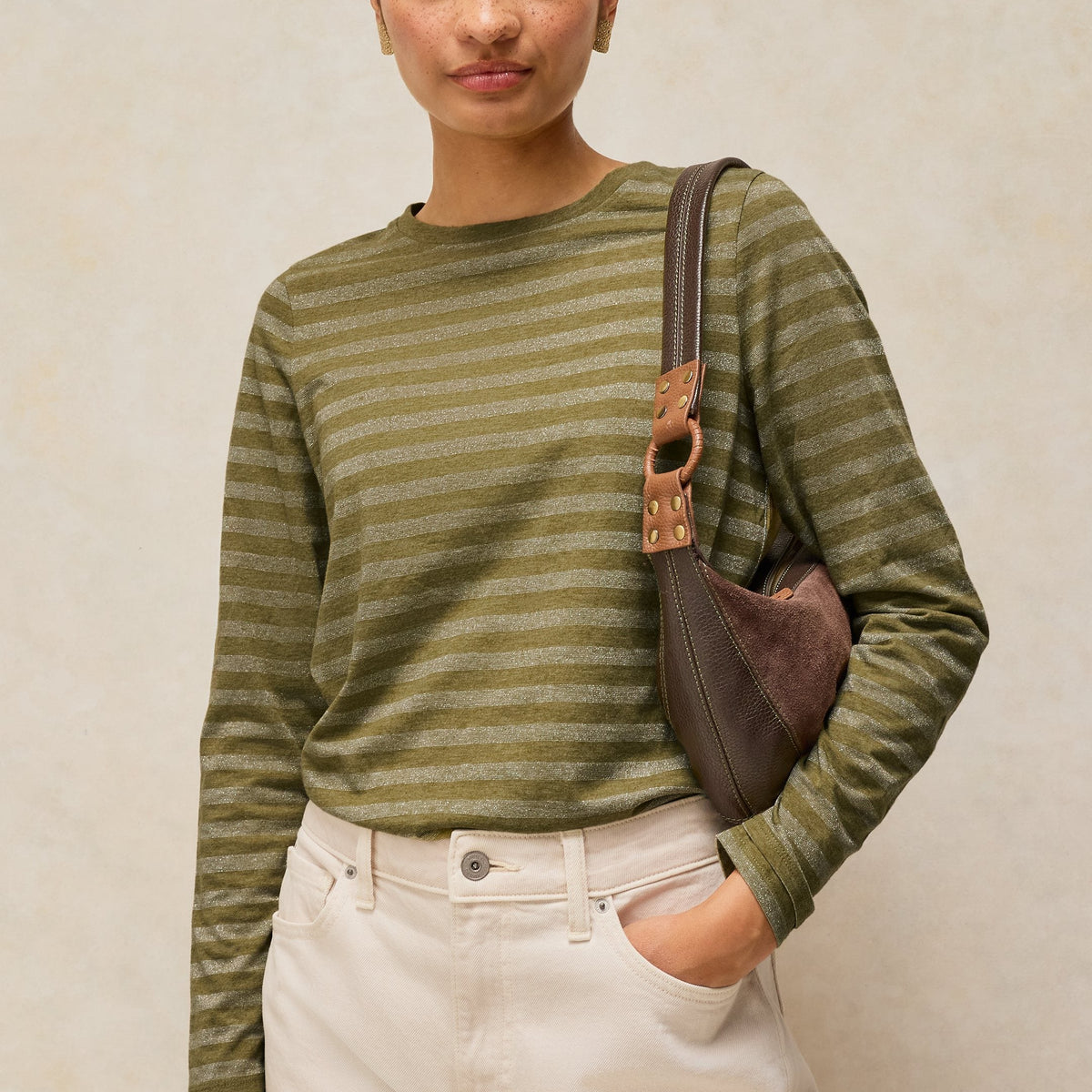 A woman wearing a green and olive striped long-sleeve knit top, cream-colored pants, and carrying a brown shoulder bag, standing against a textured beige wall.