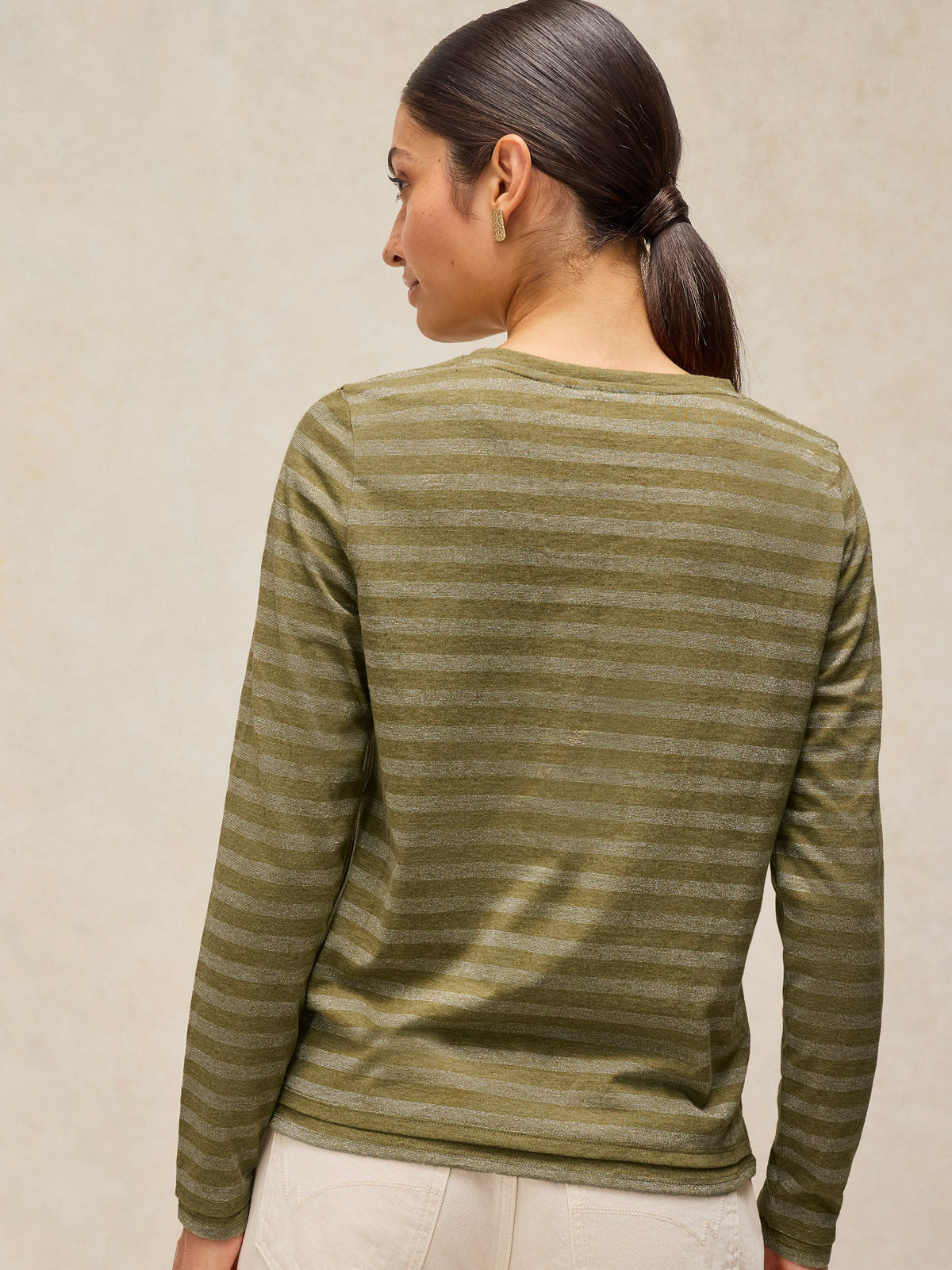 A woman shown from the back wearing a long-sleeve knit top with horizontal olive green and beige stripes, paired with off-white pants, against a textured beige background.