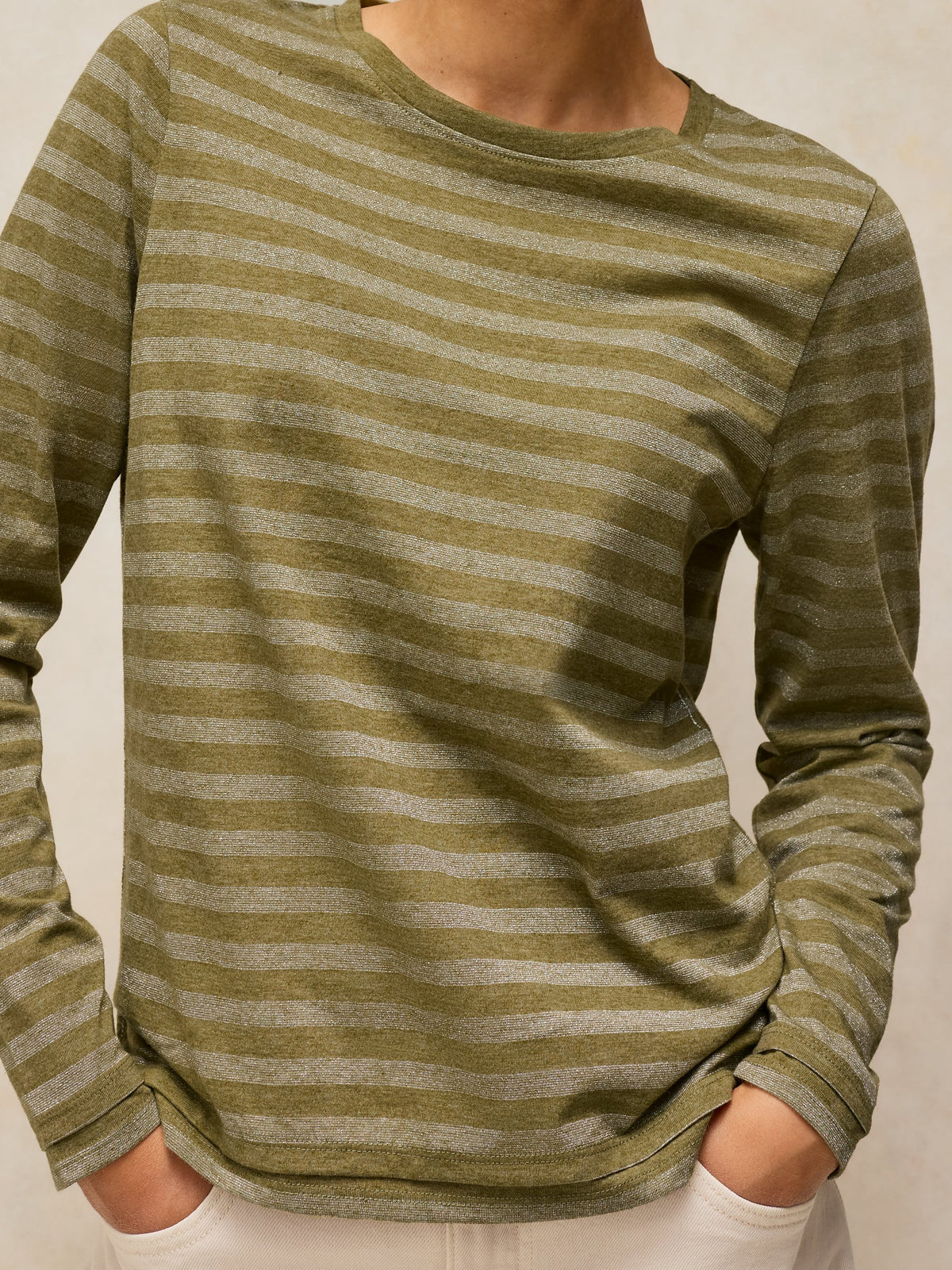 A close-up of a person wearing a long-sleeve crewneck shirt with horizontal olive green and light beige stripes, paired with white pants.