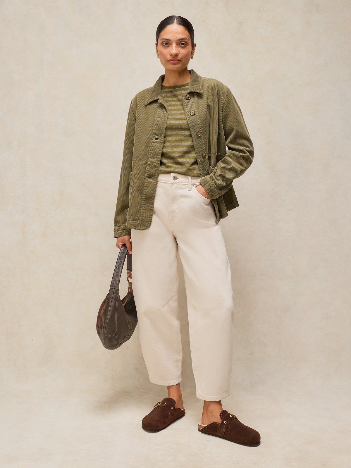 A woman stands against a textured beige studio backdrop wearing an olive green button-up overshirt over a striped green top, cream wide-leg pants, and brown mule loafers, while holding a dark brown shoulder bag.