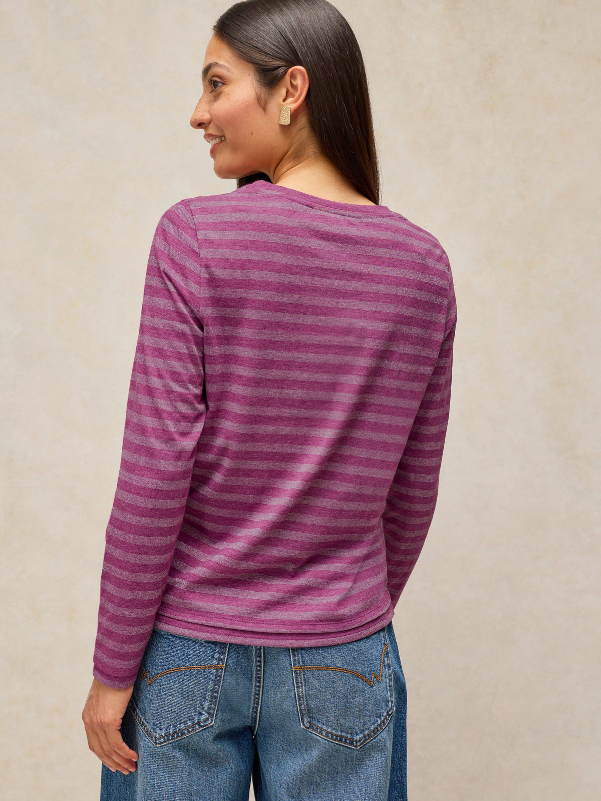 A woman shown from the back wearing a long-sleeve crewneck top with horizontal magenta and purple stripes, paired with blue jeans, against a textured beige background.