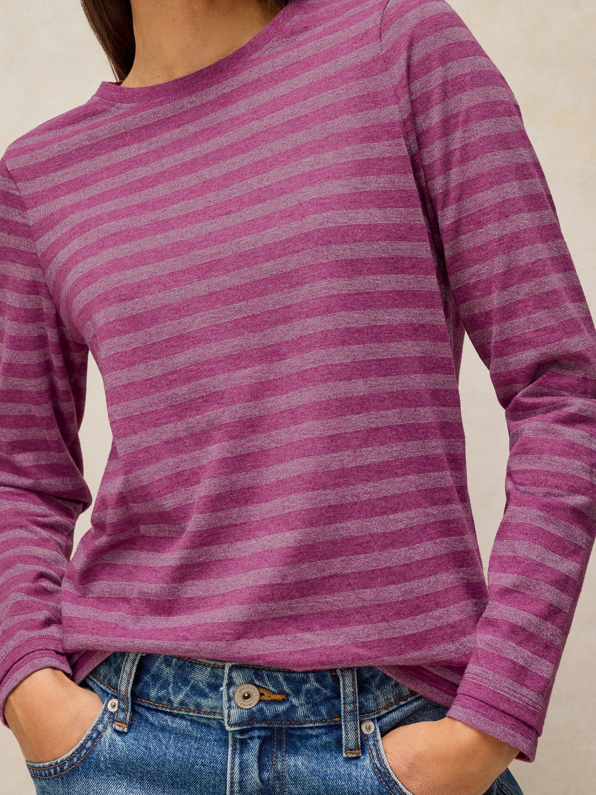 A close-up of a person wearing a long-sleeve crewneck top with horizontal magenta and dusty purple stripes, paired with blue jeans.