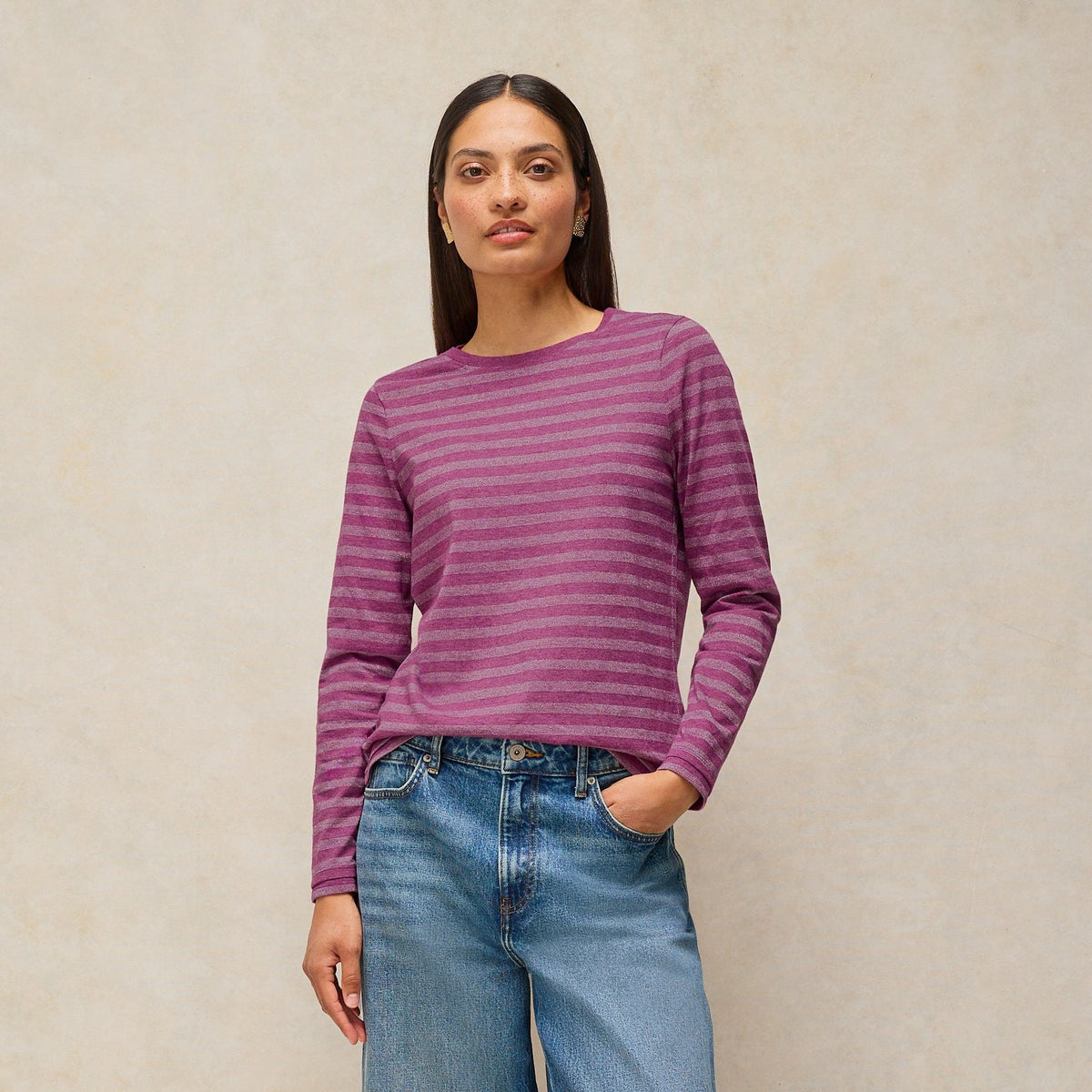 A woman stands against a textured beige studio backdrop wearing a purple and mauve striped long-sleeve knit top, wide-leg blue jeans, and brown studded loafers.