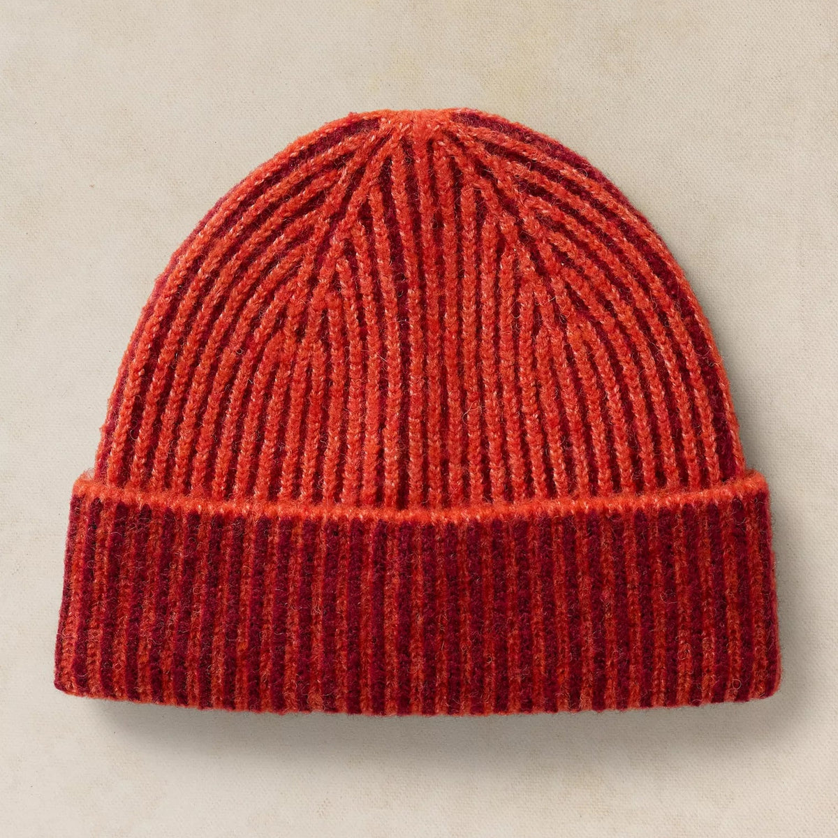 A ribbed knit beanie hat in a heathered red and orange colorway, displayed flat against a textured beige background.