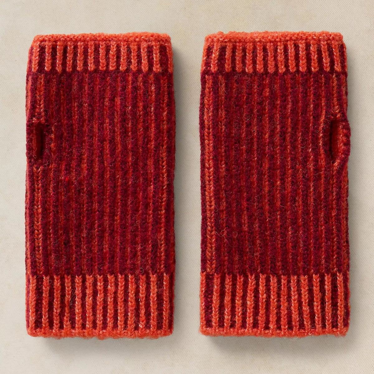 A pair of ribbed knit sleeve covers in a variegated red and orange colorway, laid flat on a light beige textured background.