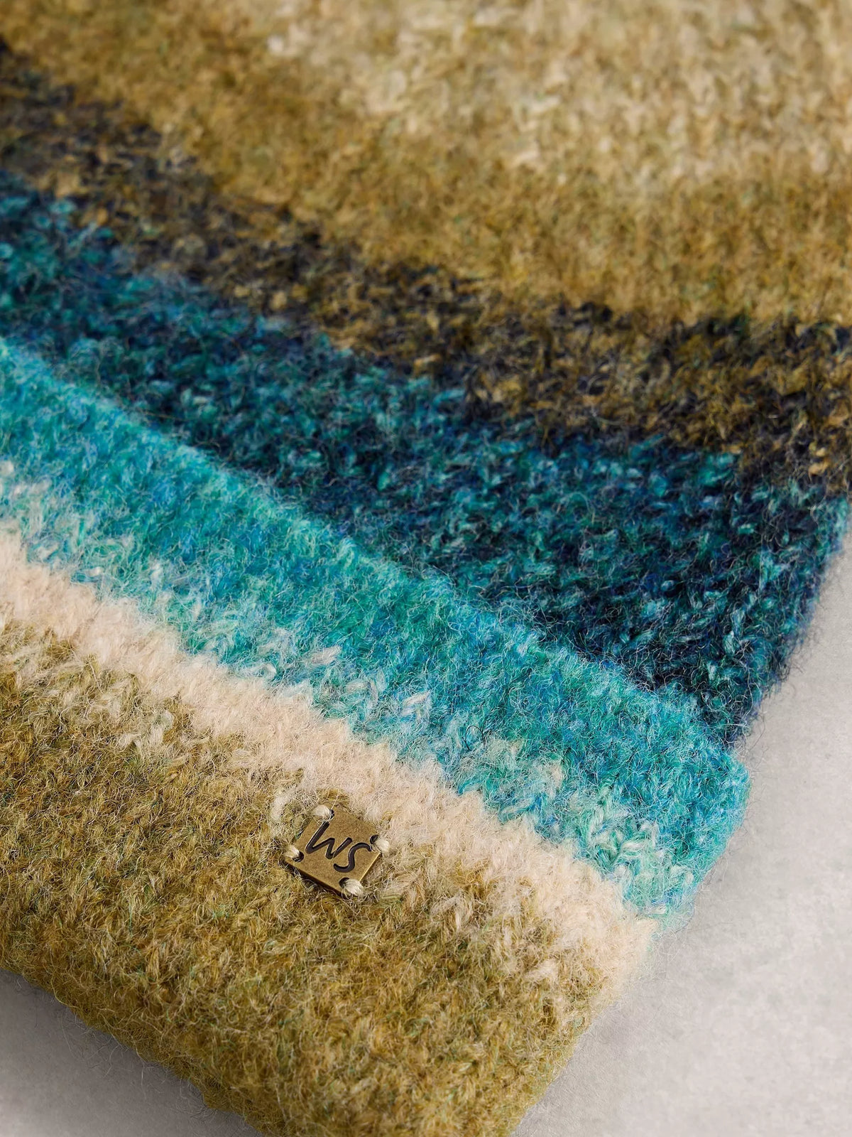 A close-up of a textured, fuzzy knit fabric with horizontal stripes in olive green, teal blue, cream, and dark navy, featuring a small metal brand tag with the initials 'ws'.
