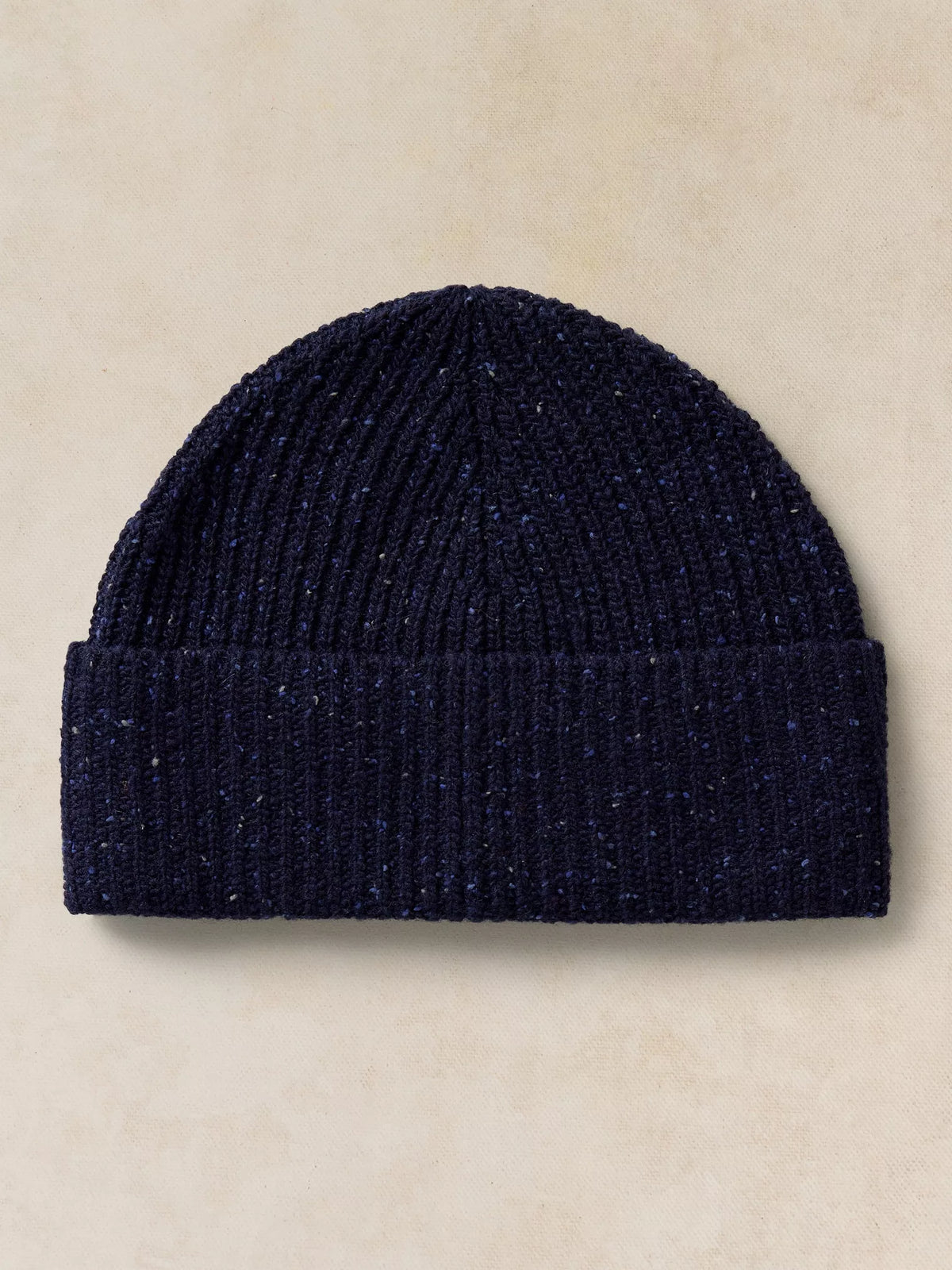 A dark navy blue ribbed knit beanie hat with subtle silver-colored flecks, presented flat against a textured beige background.