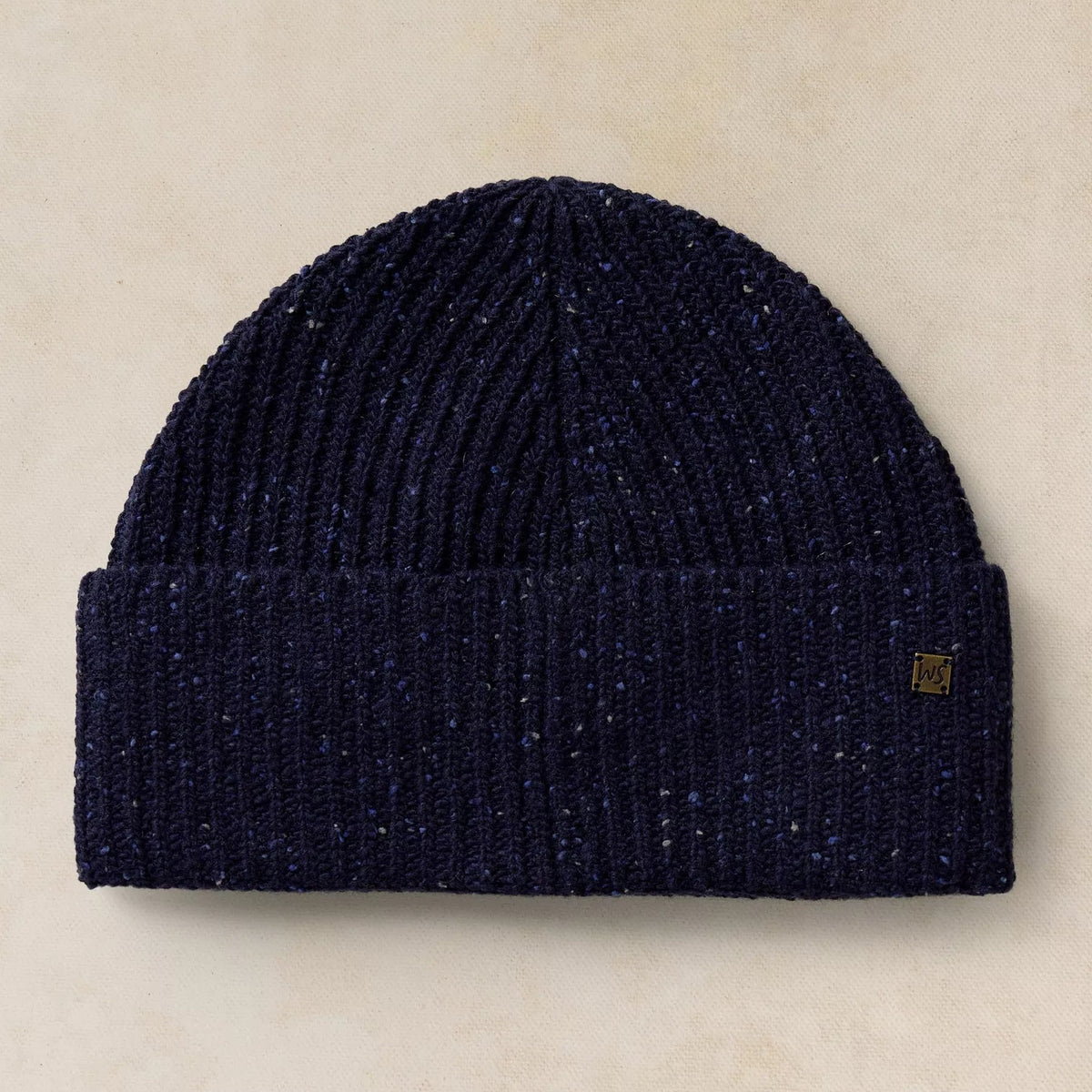 A dark navy blue ribbed knit beanie hat with subtle blue and white flecks throughout, featuring a small gold square logo patch on the cuff, set against a textured beige background.