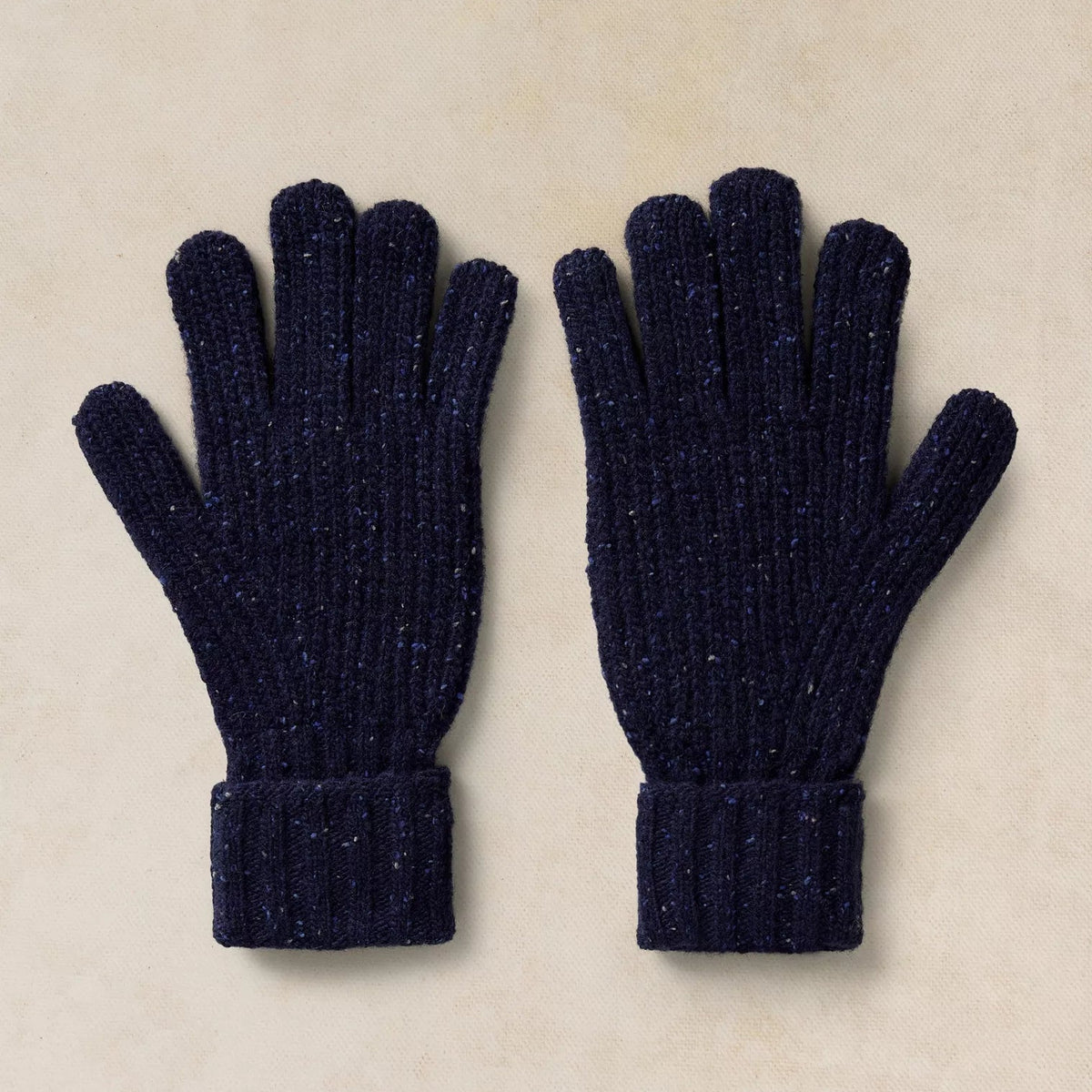 A pair of dark navy blue ribbed knit gloves with subtle glittery flecks, laid flat on a textured beige background.