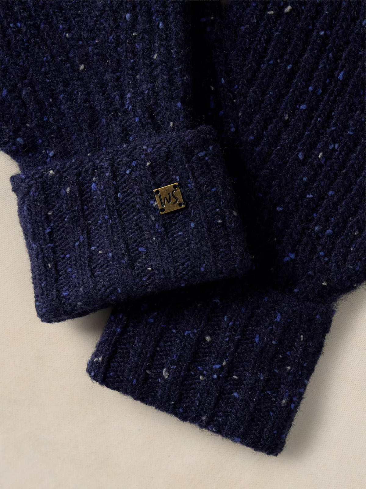 Close-up of a navy blue ribbed knit sweater with a speckled texture and a small gold square logo patch on the cuff, set against a light beige background.