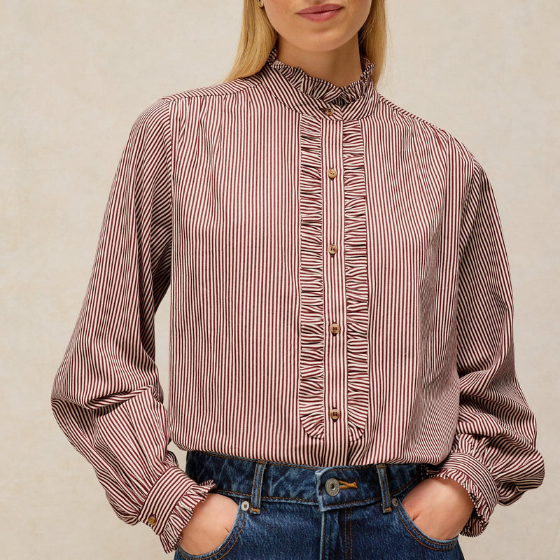 A woman wearing a long-sleeve button-front blouse with vertical stripes in dusty rose and white, paired with blue jeans, against a neutral beige background.