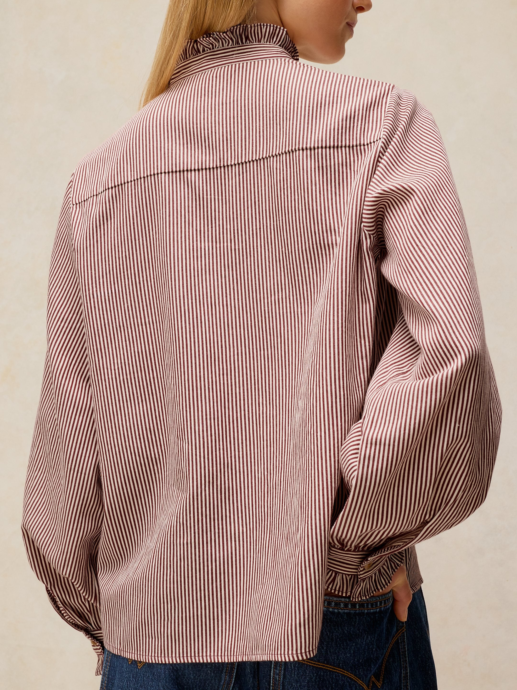 A person shown from the back wearing a long-sleeve button-up shirt with thin vertical pink and white stripes, paired with blue jeans against a plain light beige background.