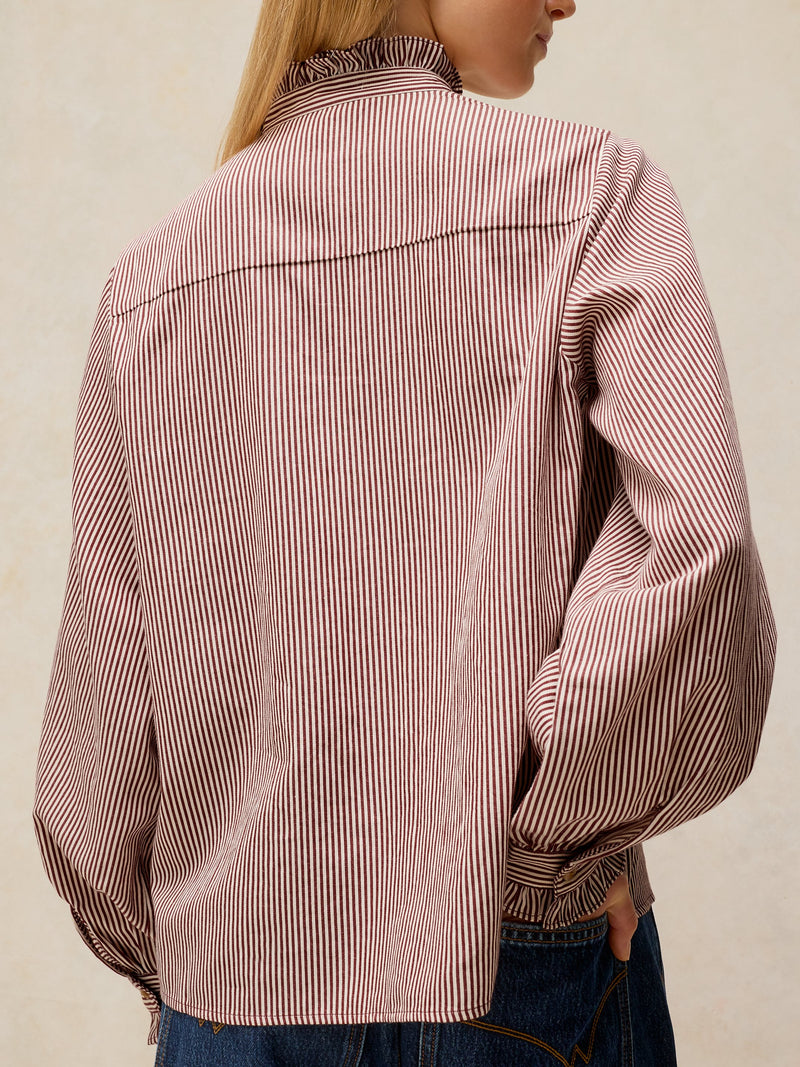 A person shown from the back wearing a long-sleeve button-up shirt with thin vertical pink and white stripes, paired with blue jeans against a plain light beige background.