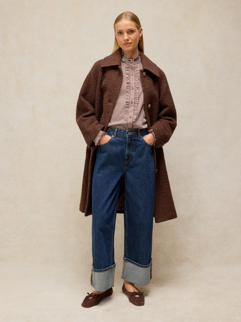 A woman stands against a neutral beige studio background wearing a dark brown textured coat over a dusty pink ruffled blouse, blue wide-leg jeans with cuffed hems, and brown loafers.