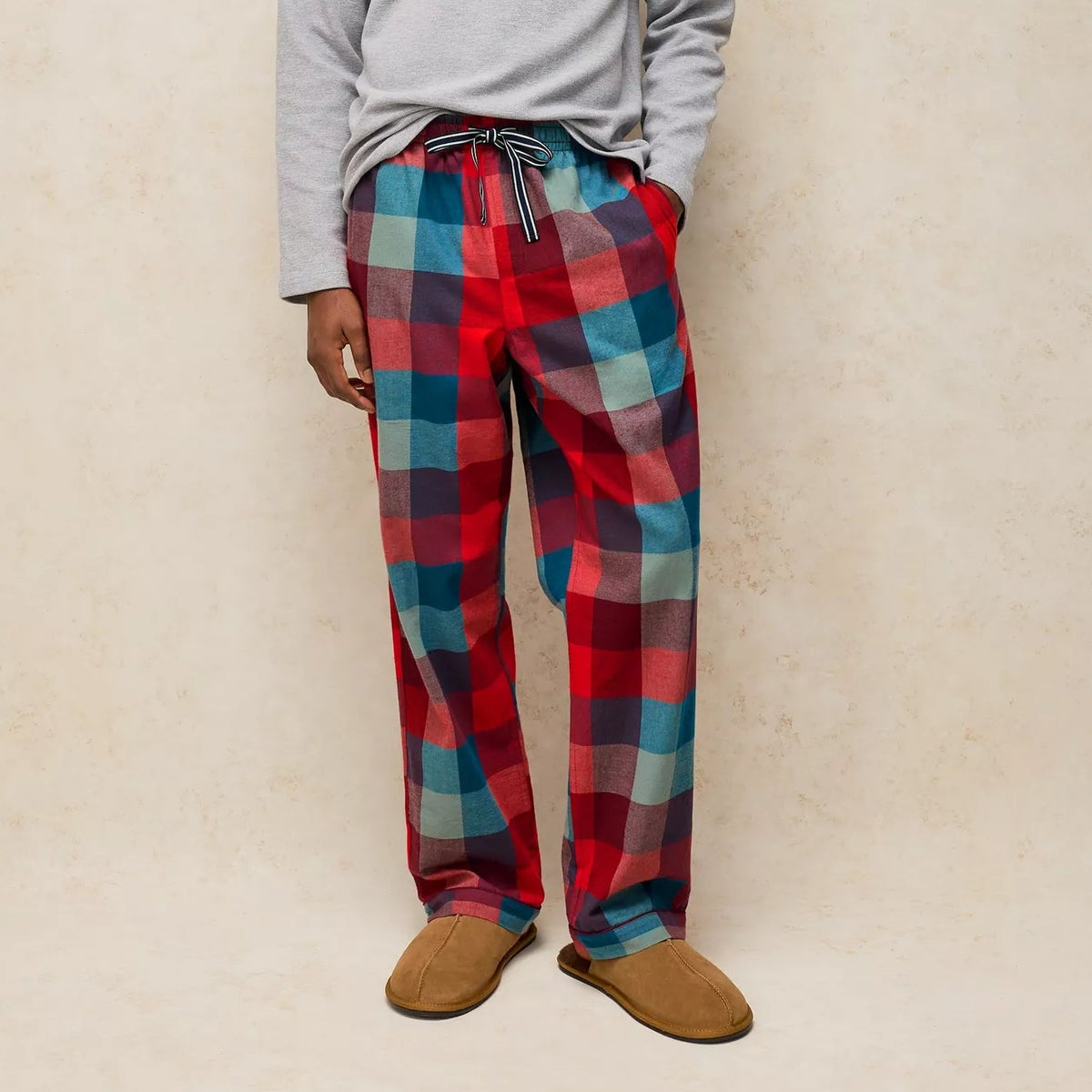 A man modeling a light gray long-sleeve henley shirt and red, blue, and purple plaid pajama pants with brown slippers, standing against a textured beige wall.