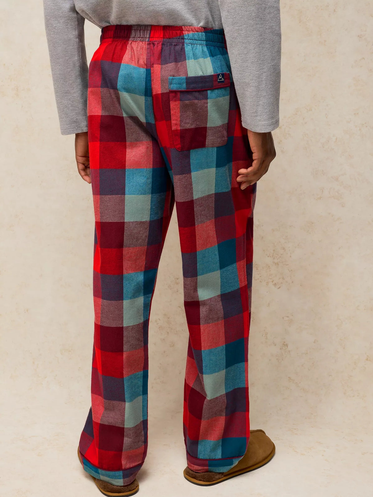 A person shown from the back wearing plaid lounge pants in red, blue, and gray, with a gray long-sleeve top and brown slippers, against a textured beige wall.