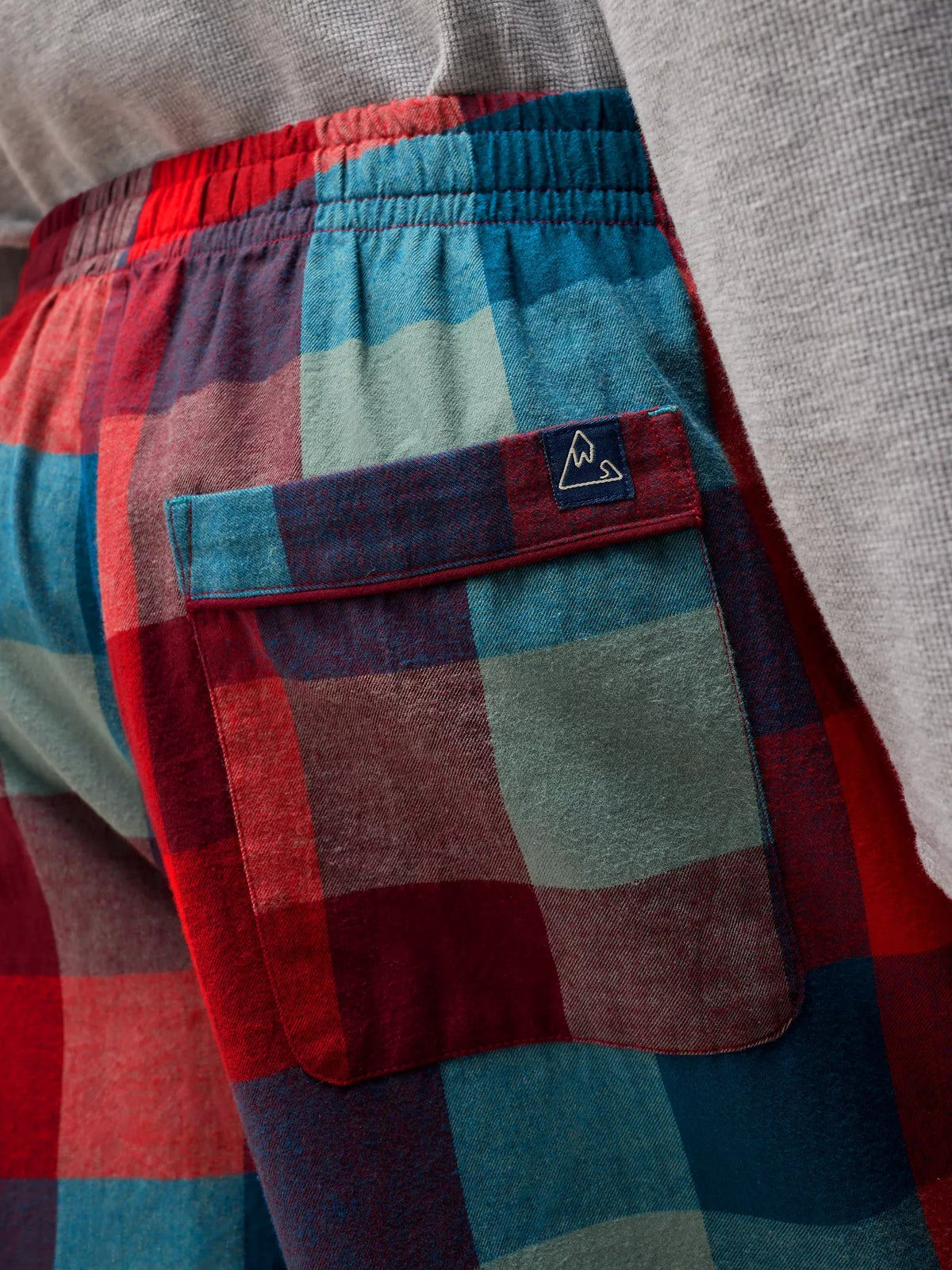 Close-up of a person wearing plaid lounge pants in red, blue, and muted green tones, with a gray knit top visible at the top and a small embroidered logo patch on the pocket.