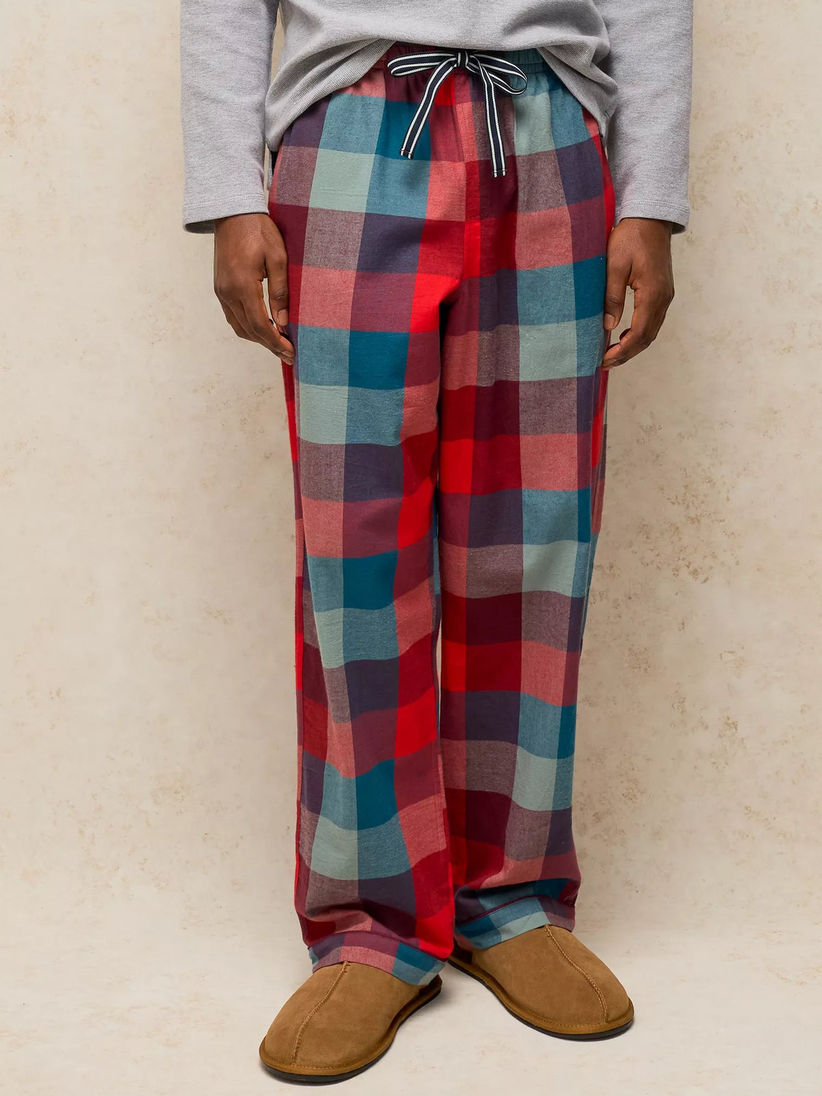 A person wearing loose plaid pajama pants in red, blue, and purple tones, paired with a gray long-sleeve top and tan slippers, standing against a textured beige wall.