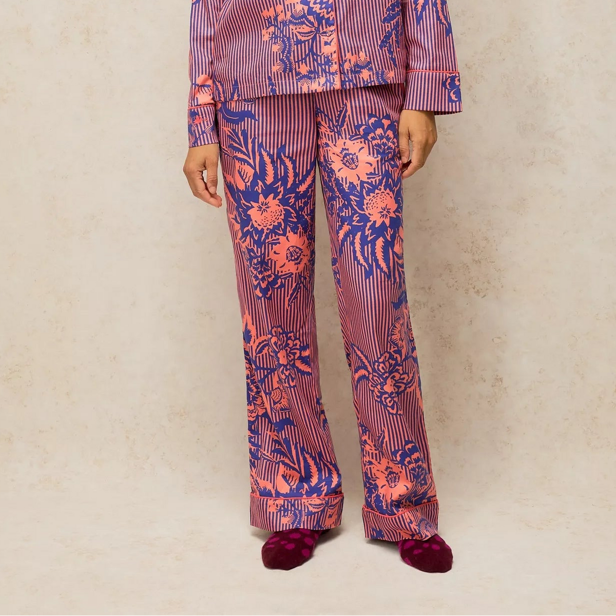 A woman modeling a matching pajama set with a purple and blue floral print over a pink striped base, standing against a textured beige wall.