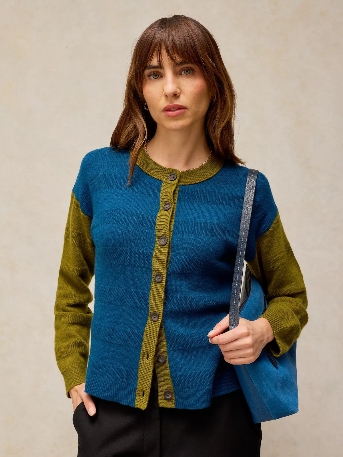 A woman wearing a color-blocked knit cardigan with a blue body and olive green sleeves and trim, holding a blue tote bag against a neutral beige background.