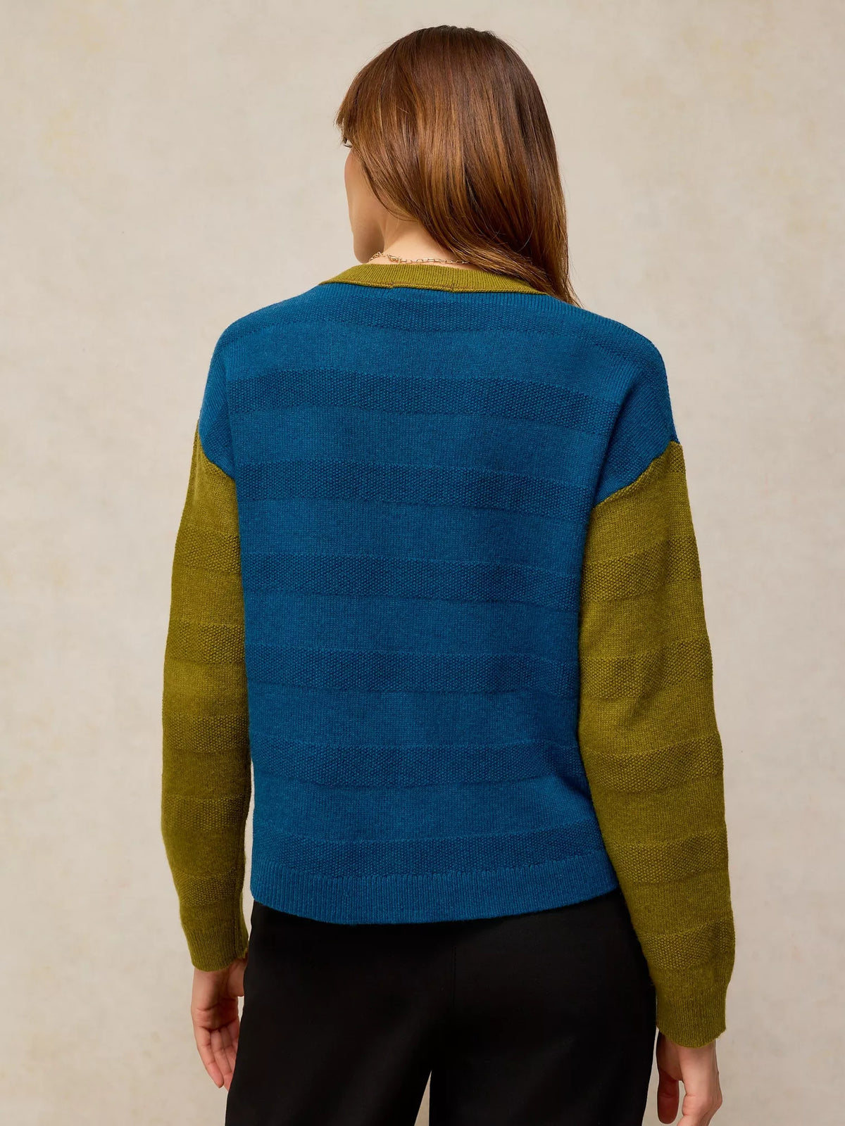 A woman viewed from the back wearing a color-block knit sweater with a blue body and olive green sleeves, paired with black pants, against a textured beige background.