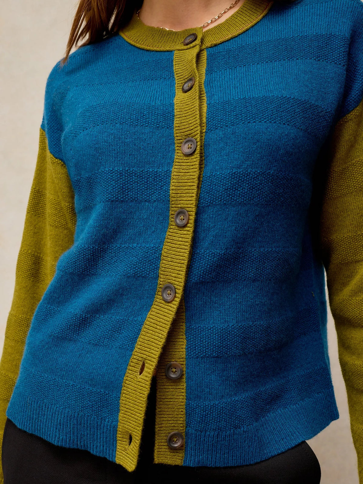 A close-up of a person wearing a knit cardigan sweater with horizontal blue and teal stripes, olive green trim, and dark buttons.