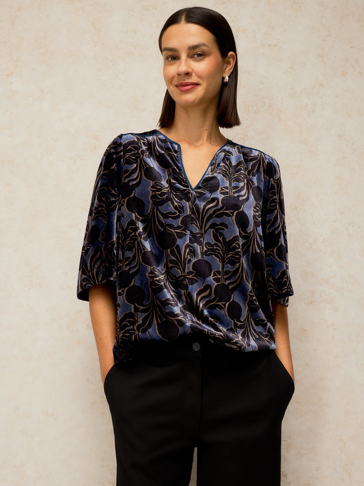 A woman wearing a dark blue and black patterned blouse with three-quarter sleeves and black trousers, standing against a textured beige wall.
