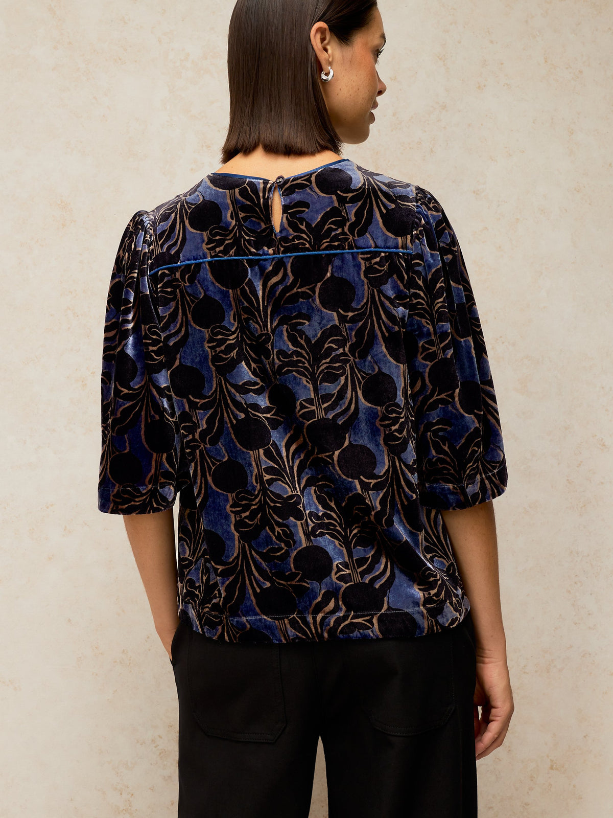 A woman viewed from the back wearing a dark blue velvet blouse with a black and gold floral pattern, paired with black pants, against a textured beige wall.