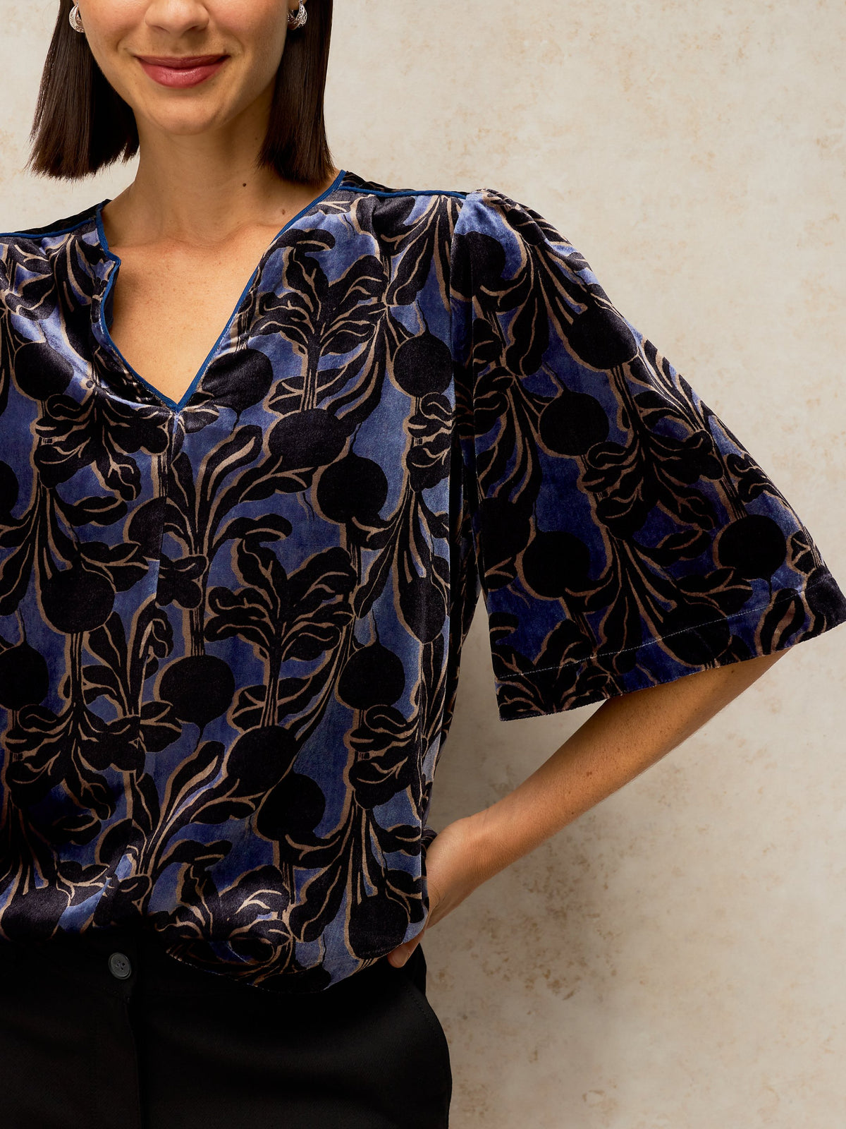 A woman wearing a blue and black patterned blouse with short flutter sleeves and a V-neckline, paired with black trousers, standing against a textured beige wall.