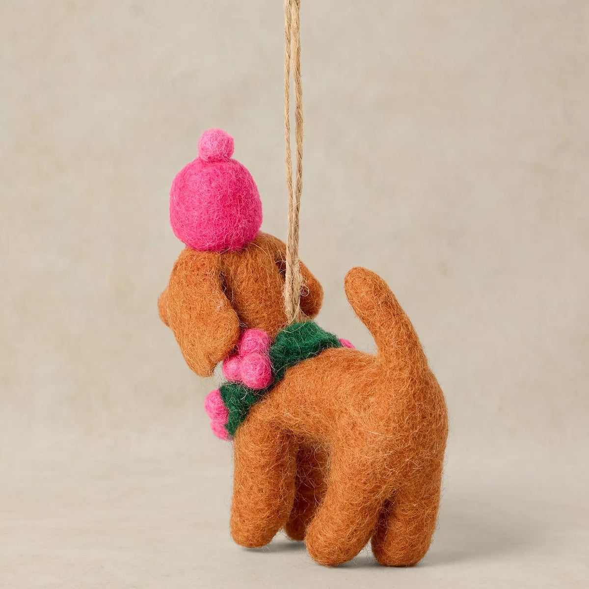 A felted wool dog ornament shown from the back, crafted in a warm brown with a bright pink hat, a green scarf, and pink pom-poms, hanging by a natural jute string against a neutral beige background.