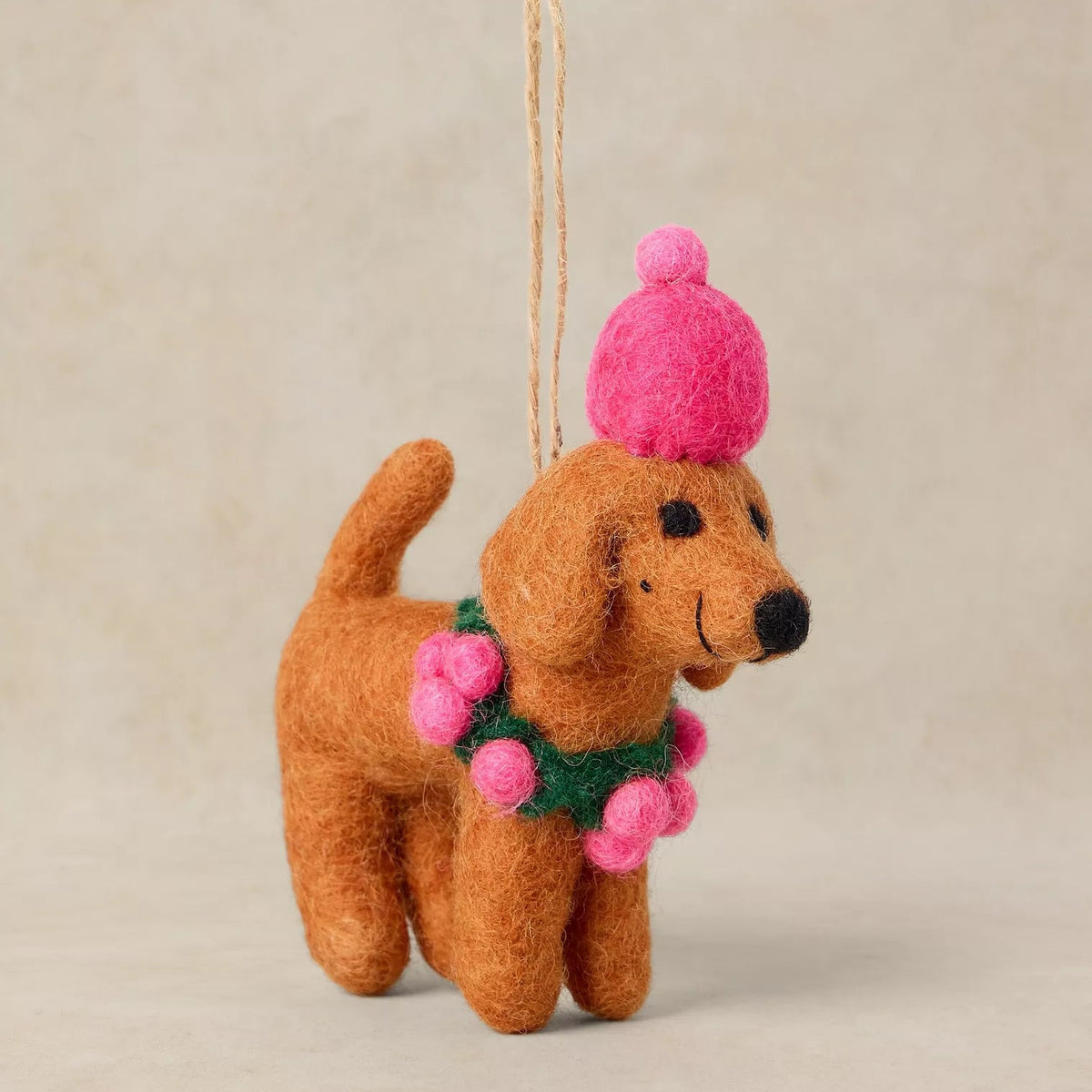 A felt dog ornament in warm brown with a bright pink hat, pink pom-poms, and a green collar, hanging by a string against a neutral beige background.
