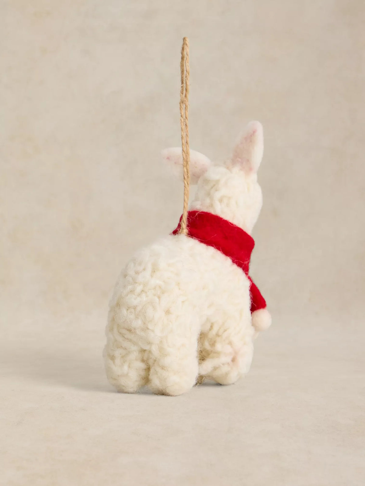A small white plush alpaca or llama ornament viewed from the back, wearing a bright red scarf and hanging from a natural jute string against a warm beige studio background.