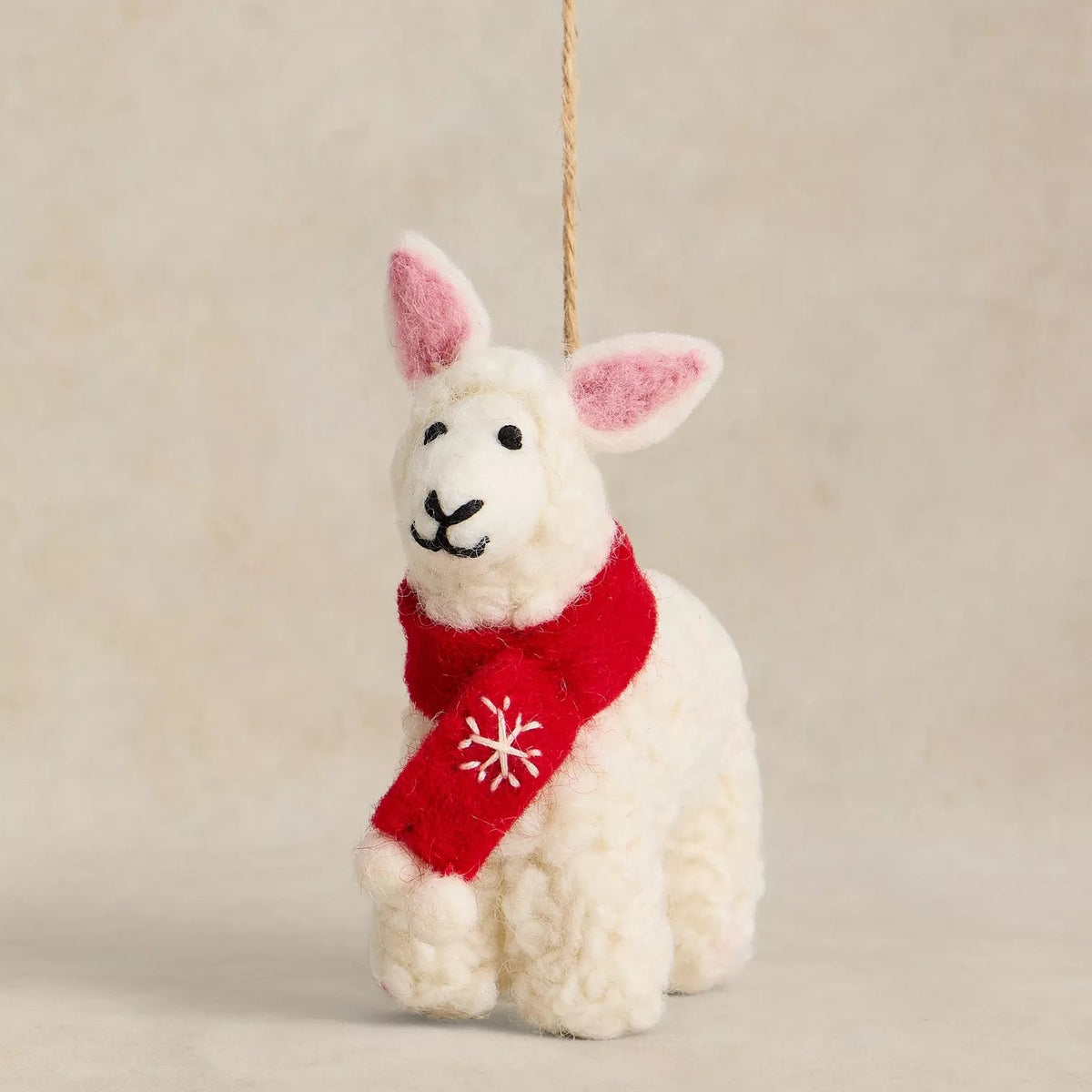 A small white felted wool sheep ornament with pink inner ears, wearing a red knit scarf with a white snowflake, hanging against a warm beige background.