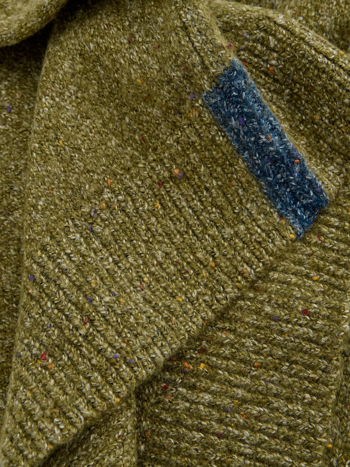 Close-up of a textured tweed knit garment in olive green with multicolored flecks and a contrasting blue fabric tab near the collar.