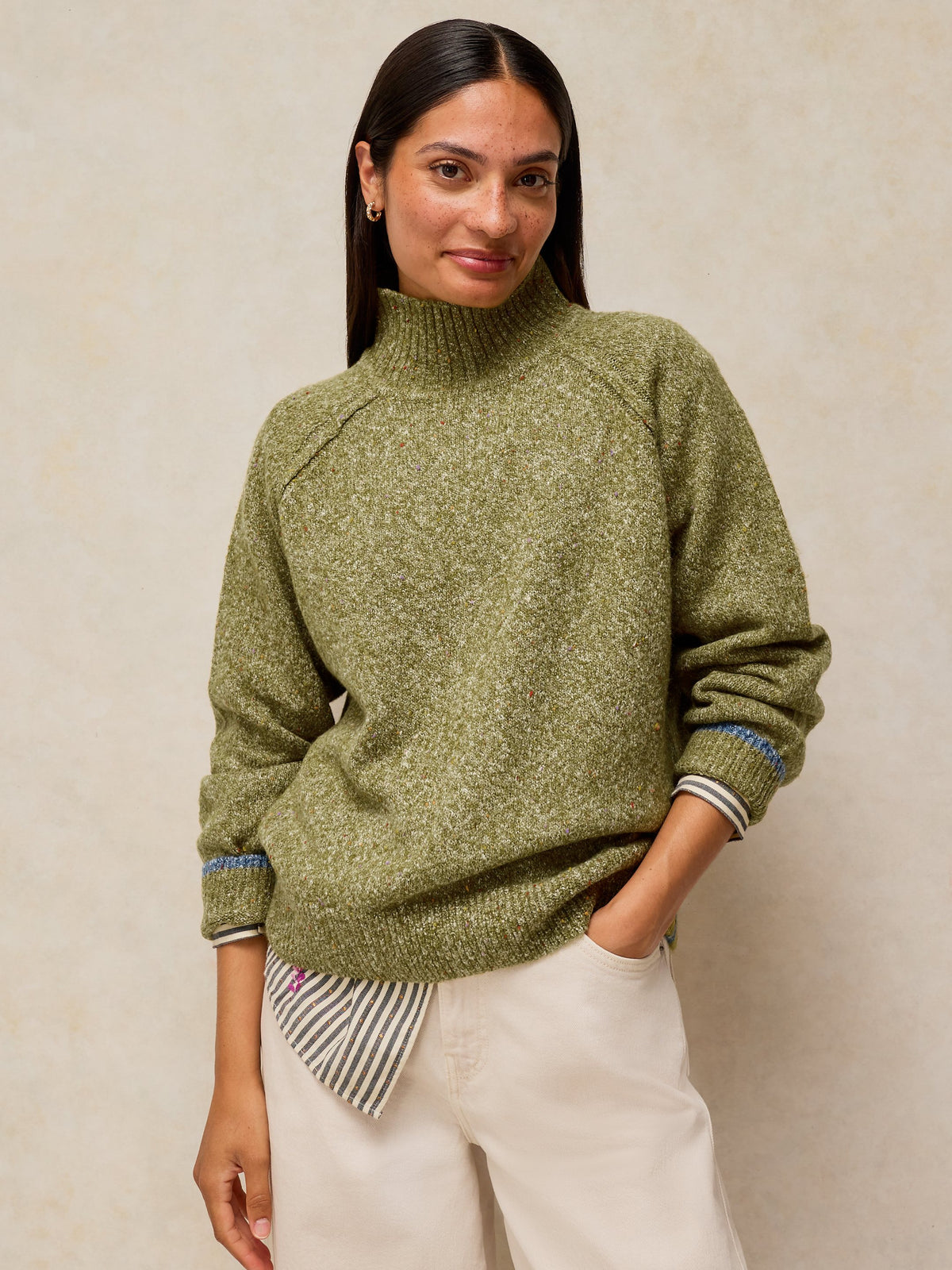 A woman wearing a textured olive green mock-neck sweater with striped ribbed cuffs, paired with cream-colored pants, standing against a neutral beige wall.