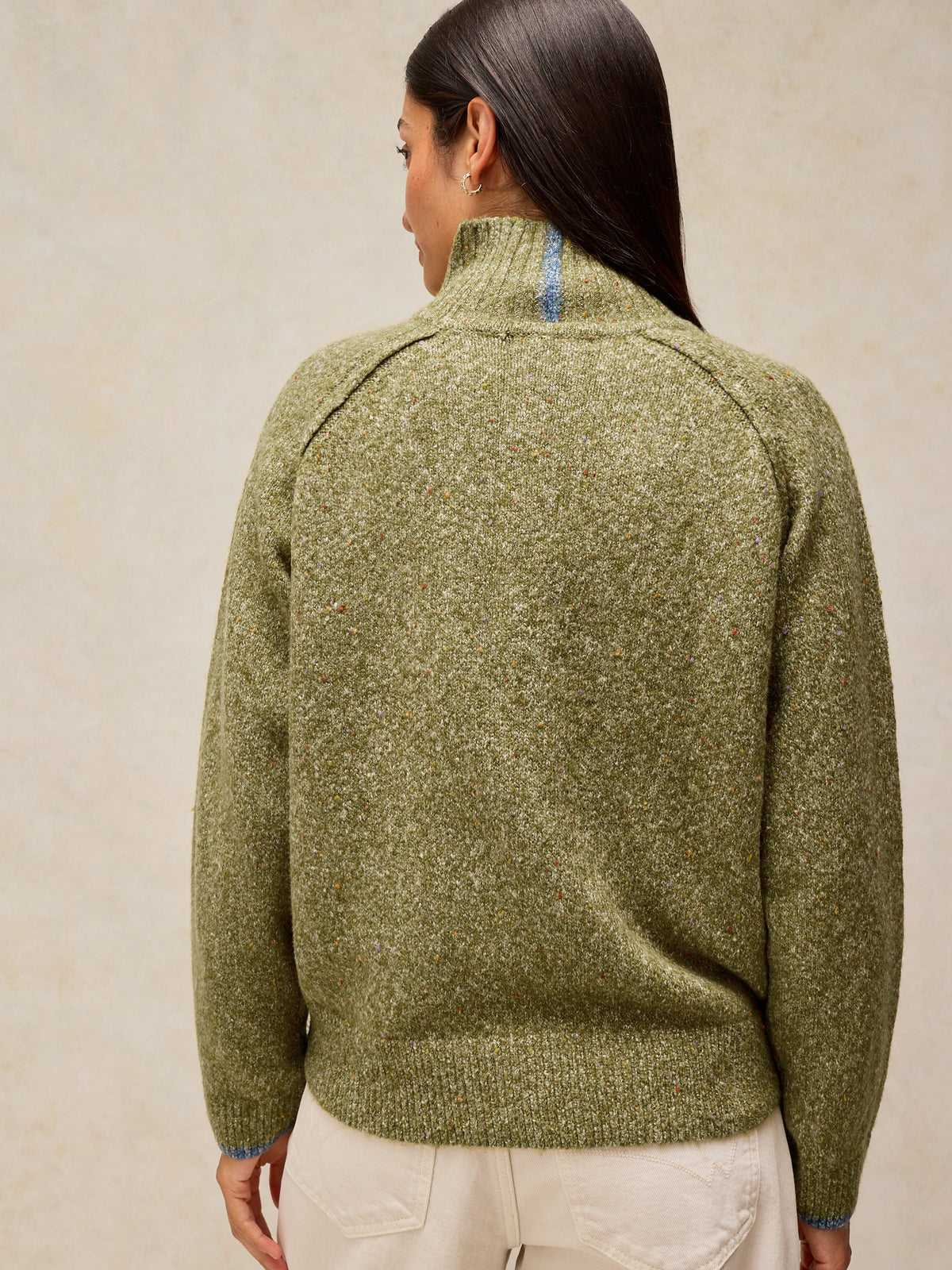 A woman shown from the back wearing a textured olive green mock-neck sweater and cream pants against a neutral beige background.
