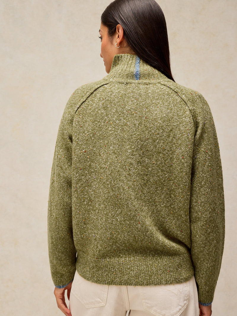 A woman shown from the back wearing a textured olive green mock-neck sweater and cream pants against a neutral beige background.