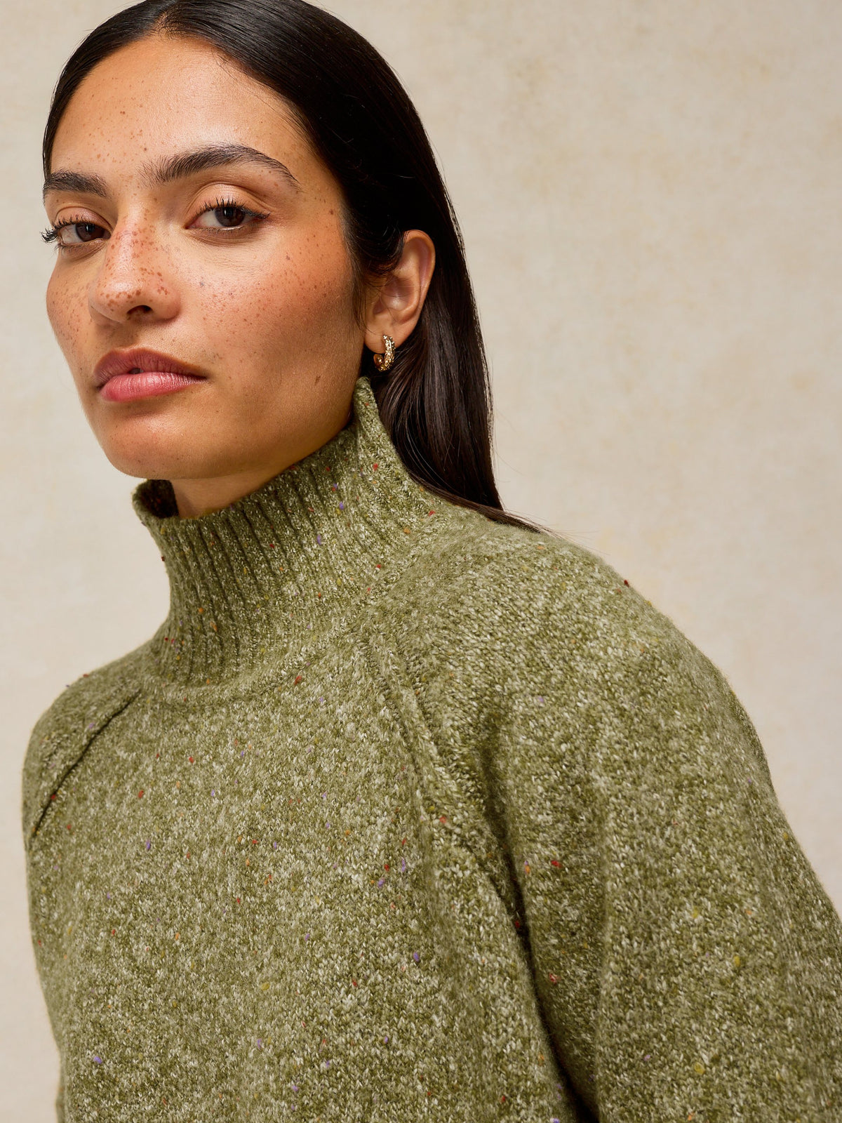 A close-up of a woman wearing a textured olive green turtleneck sweater against a neutral beige background.