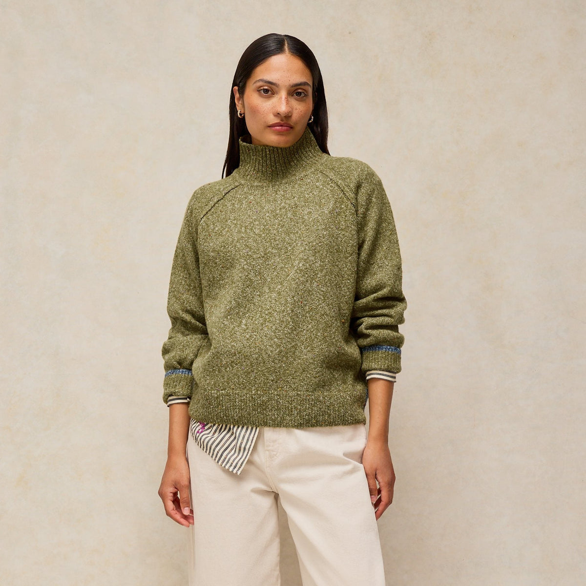 A woman stands against a textured beige studio backdrop wearing an olive green knit mock-neck sweater, off-white wide-leg trousers, and burgundy flats.