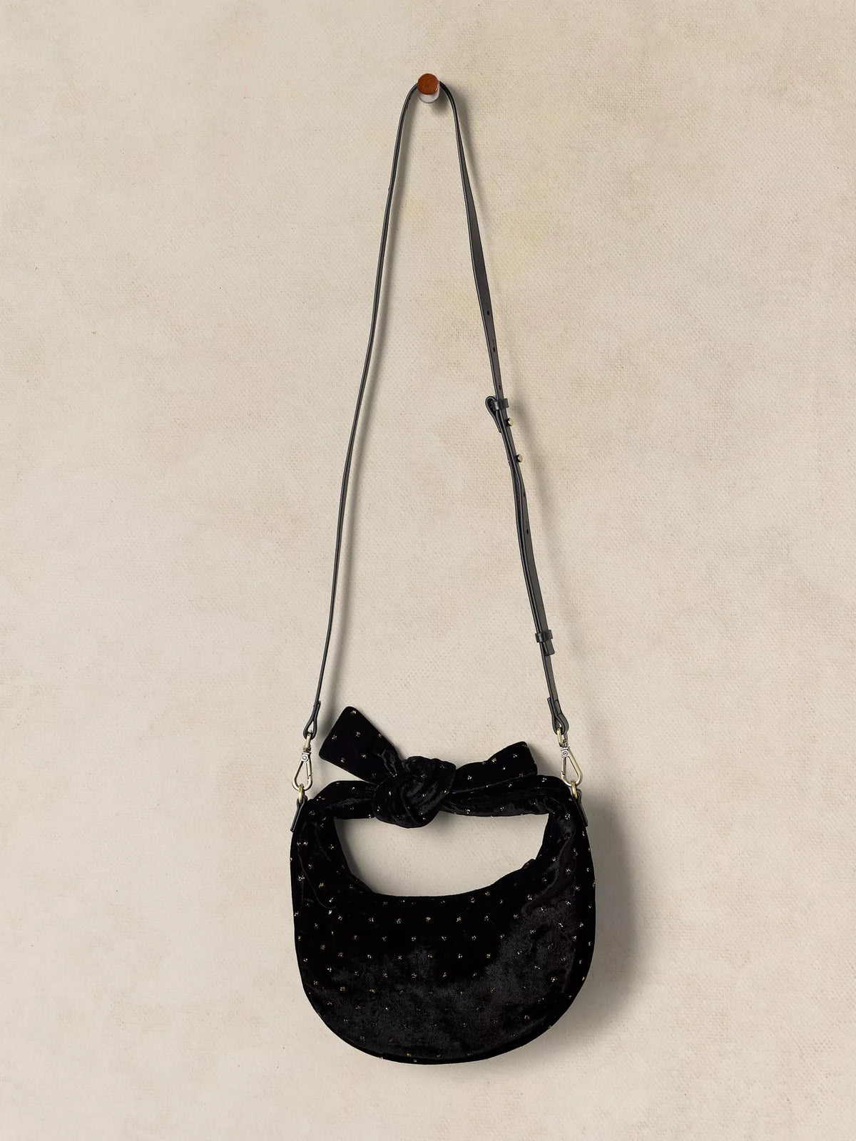 A black velvet crescent-shaped shoulder bag with a knotted bow detail at the top and small metallic studs, hanging from a hook against a textured beige wall.