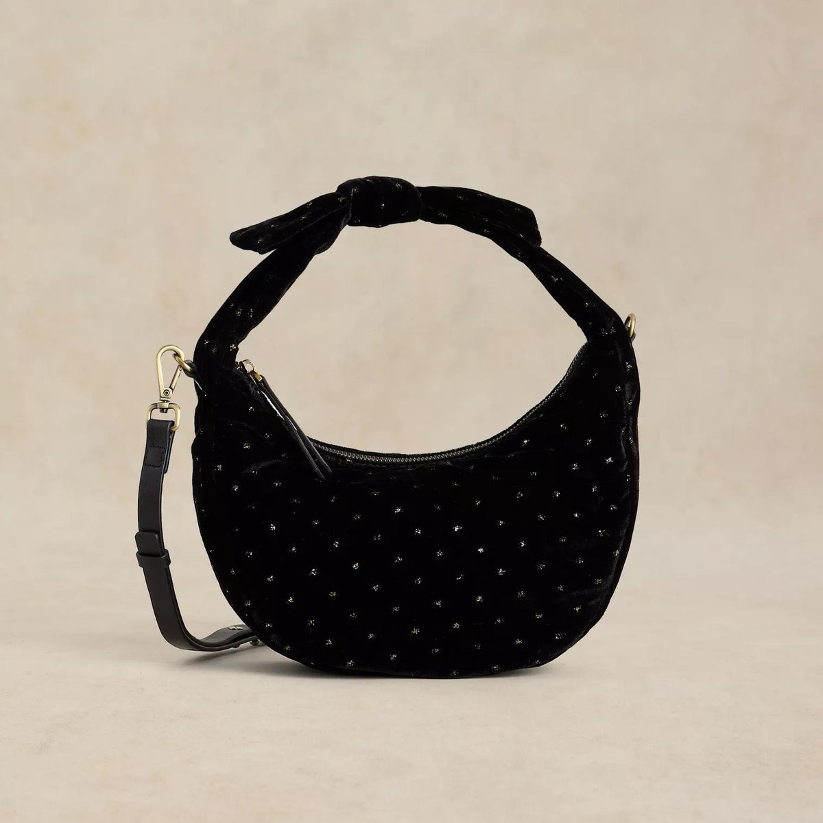 A black velvet crescent-shaped shoulder bag with silver rhinestone embellishments and a knotted top handle, shown against a neutral beige textured background.