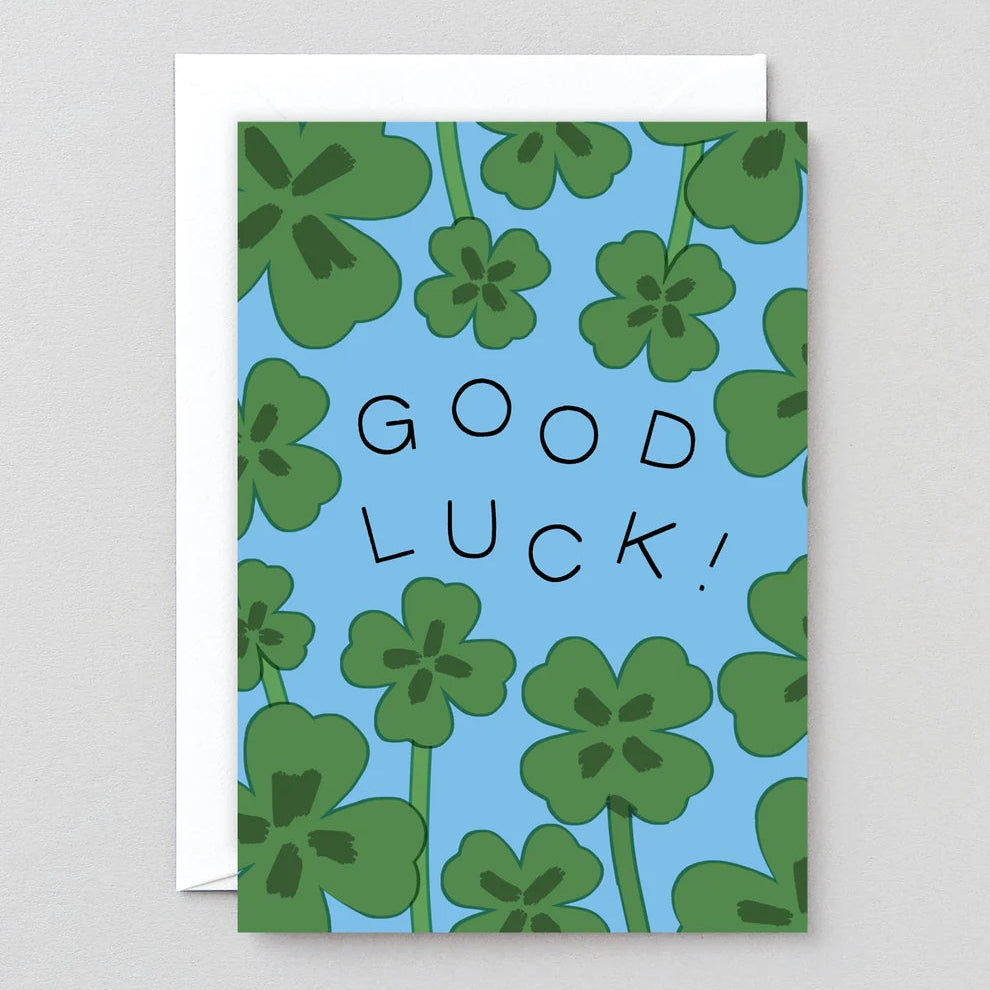 A square greeting card with a light blue background, green shamrock pattern, and the words 'GOOD LUCK!' in black hand-drawn letters, shown with a white envelope behind it on a light gray surface.