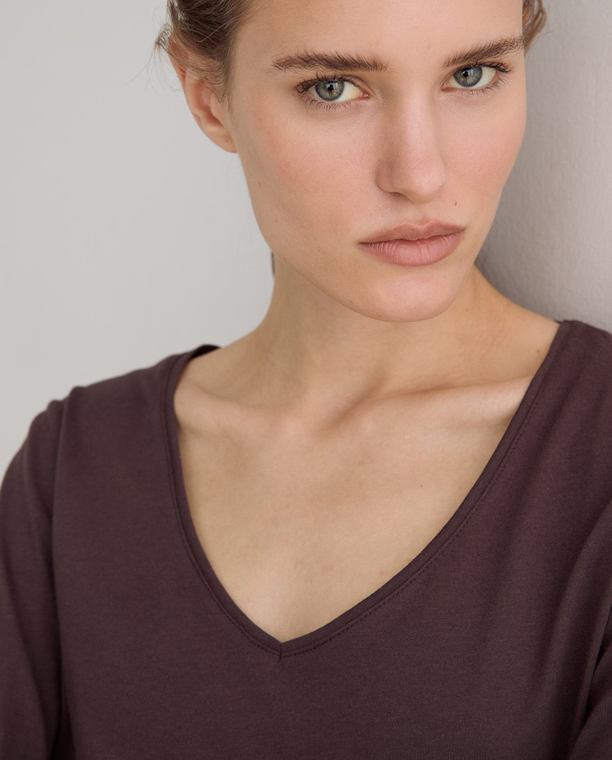 Close-up of a woman wearing a dark plum purple long-sleeve V-neck top against a light gray background.