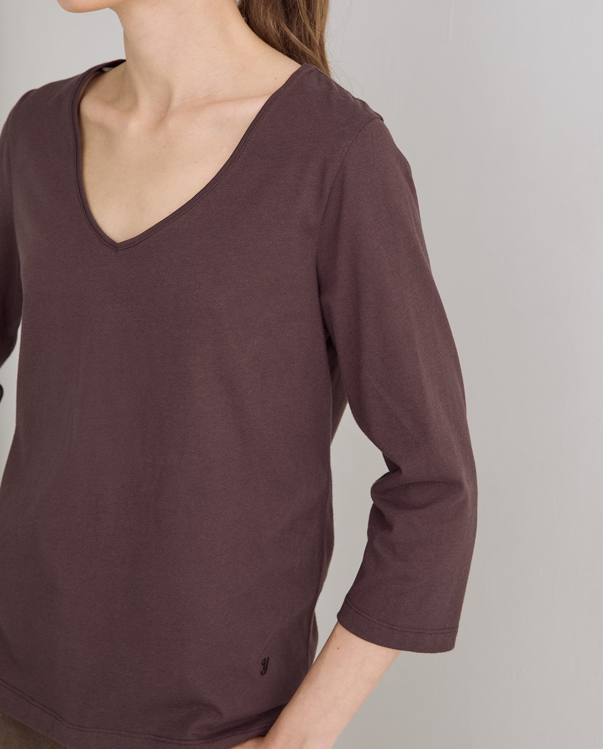 A close-up of a woman wearing a dark brownish-purple v-neck long-sleeve top, showing the fabric texture and 3/4 sleeve length against a neutral gray background.