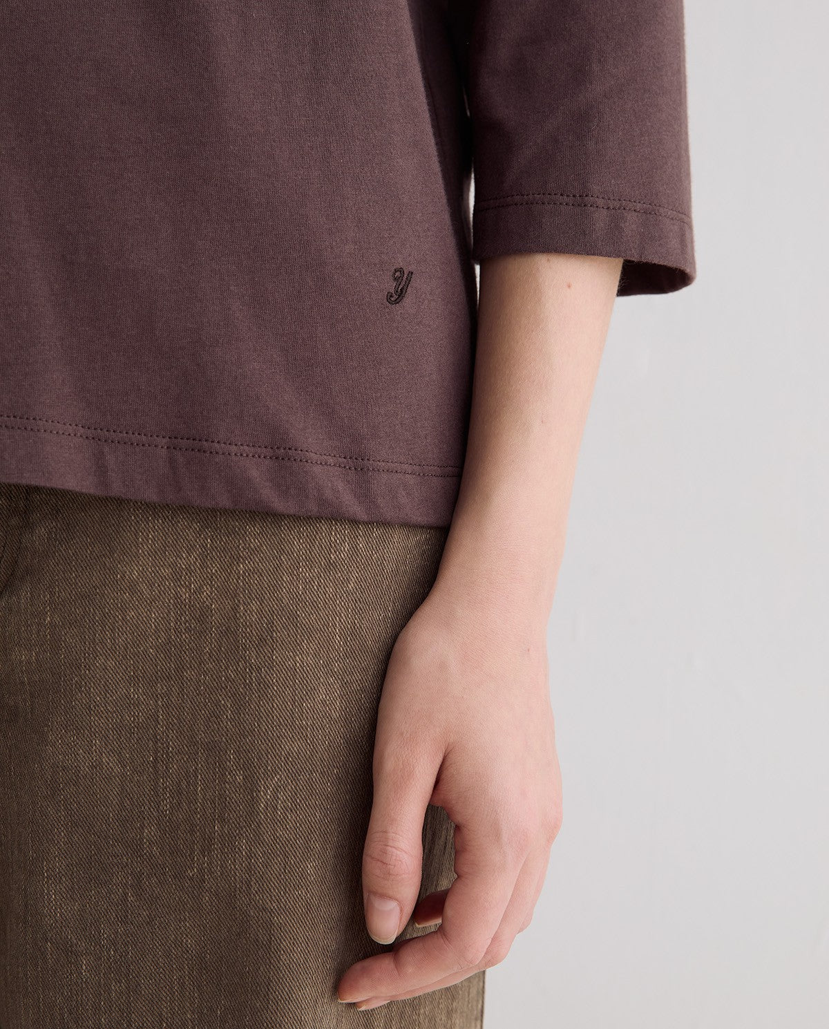 Close-up of a person wearing a muted mauve-brown short-sleeve top and textured brown bottoms, with the hand resting near the waist against a plain light gray background.