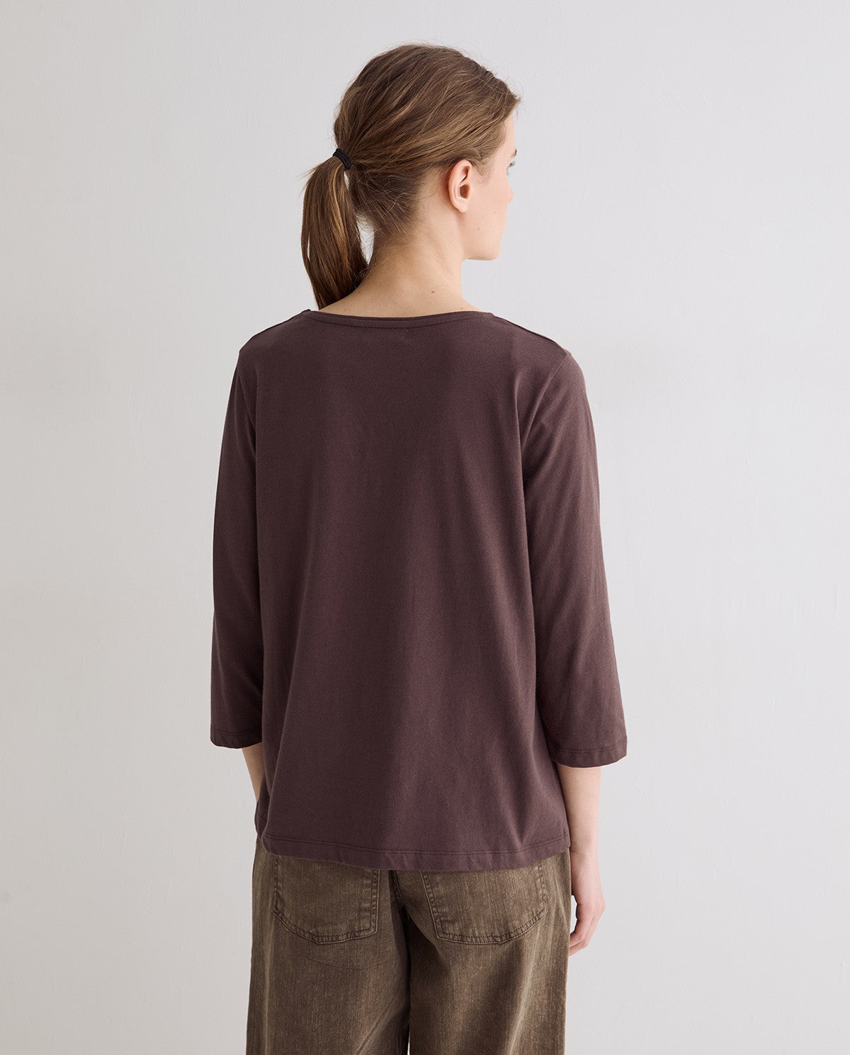 A woman viewed from the back wearing a dark brown, three-quarter sleeve crewneck top and brown pants, set against a plain light gray studio background.