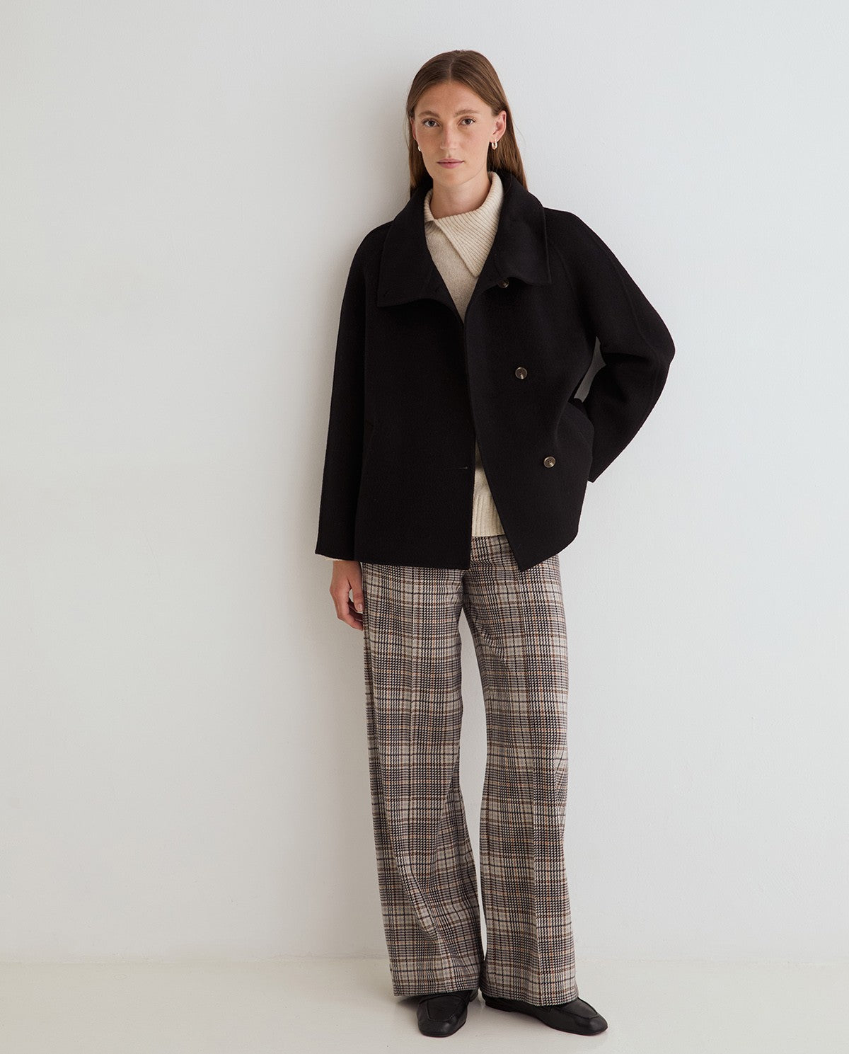 A model stands against a plain light gray studio wall wearing a black double-breasted coat over a cream turtleneck, paired with brown and beige plaid wide-leg trousers and black shoes.