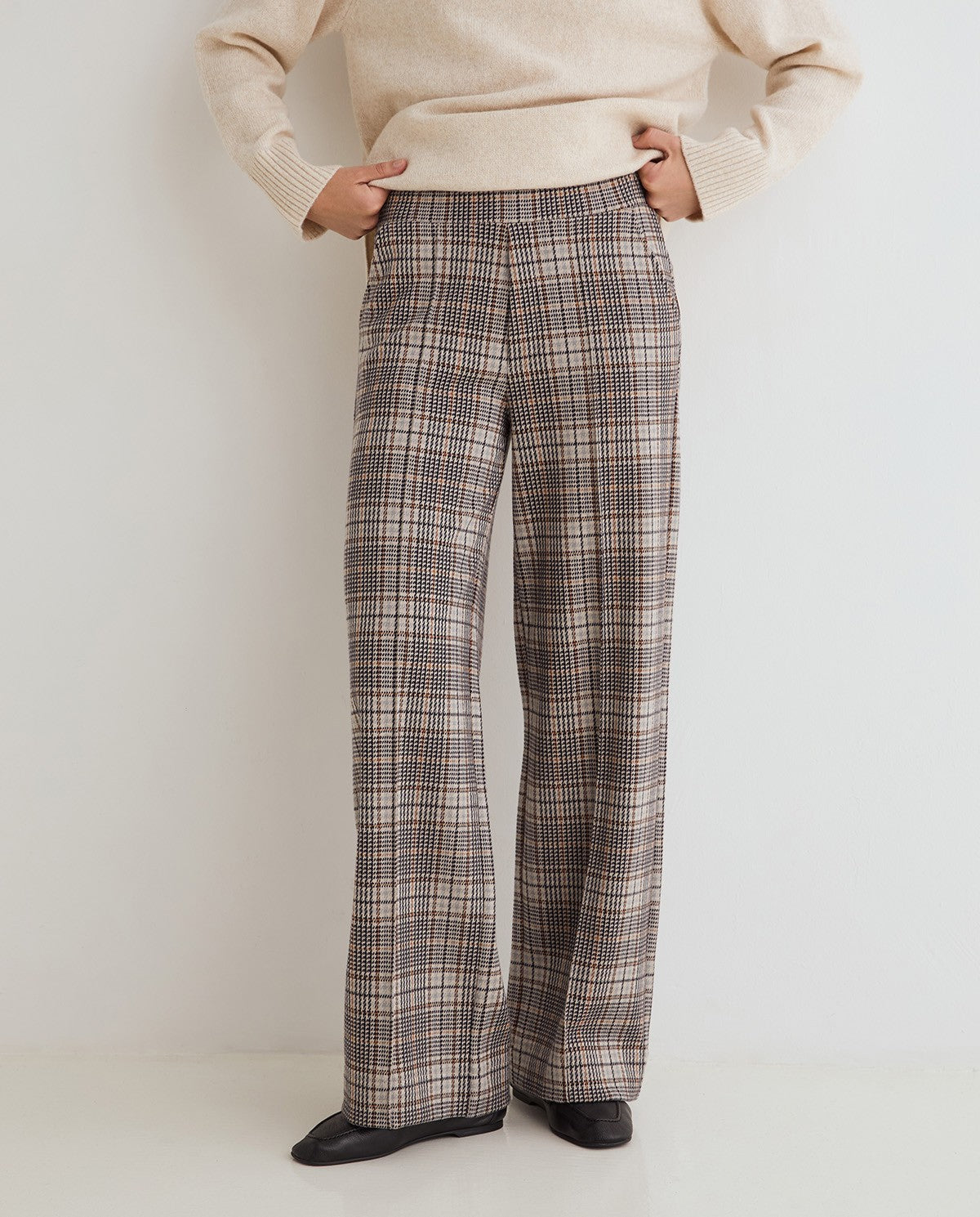 A person wearing high-waisted, wide-leg plaid trousers in a brown, beige, and gray pattern, paired with a cream sweater and black shoes, standing against a plain white studio background.